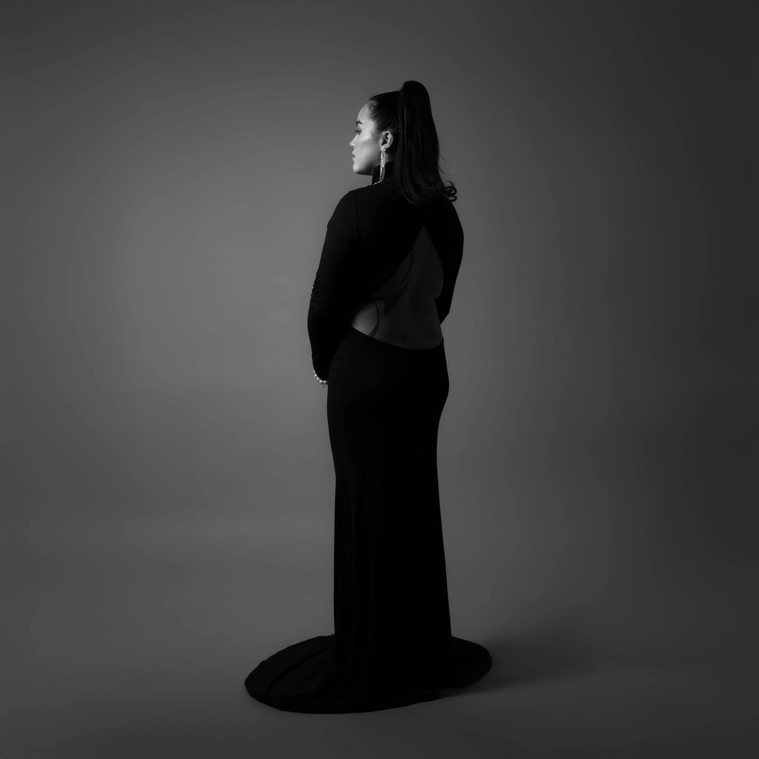 A woman in a long black dress with a sheer back, standing sideways against a gradient gray background.