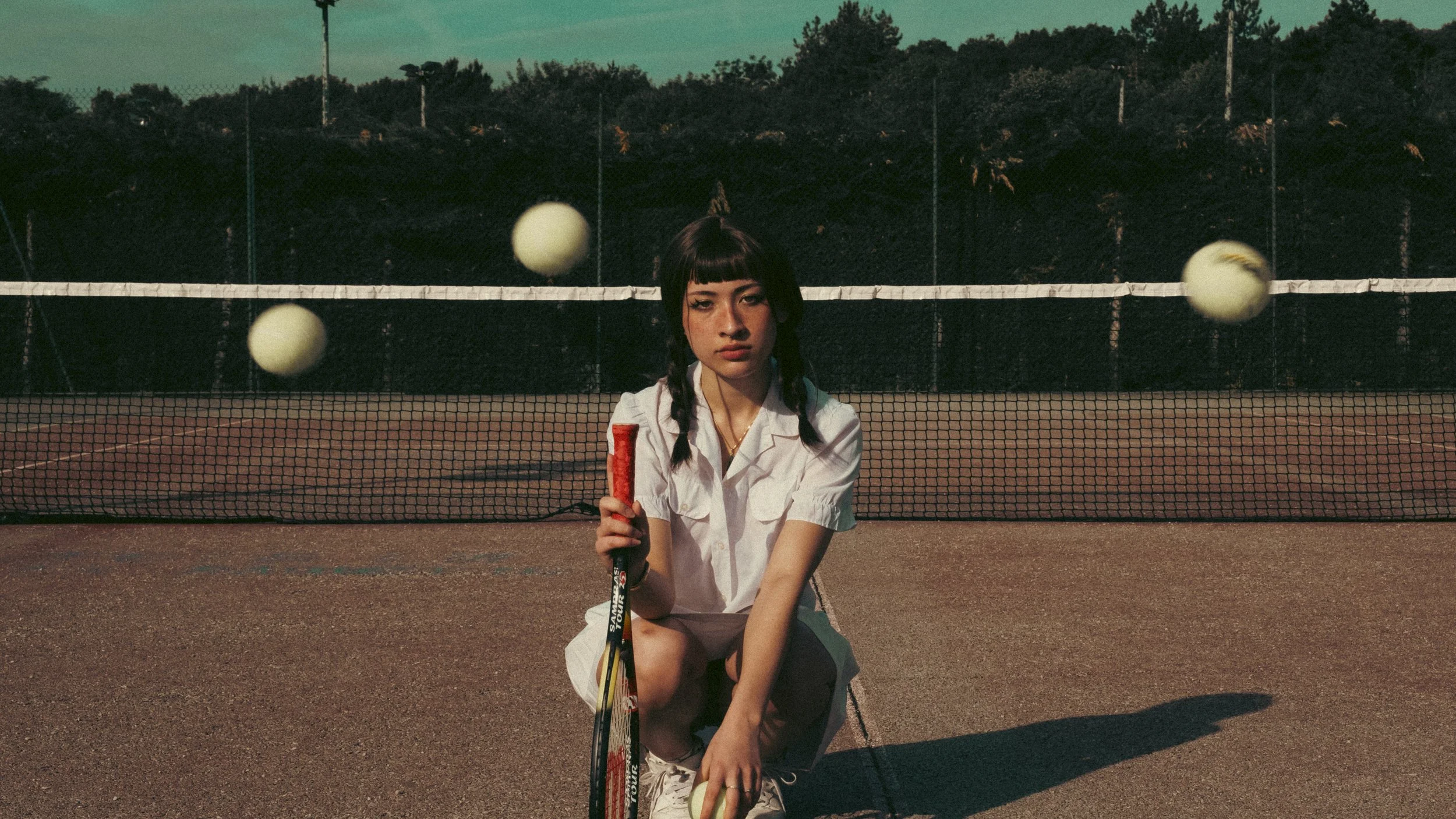 A young woman in a white tennis outfit crouches on a tennis court holding a tennis racket and a tennis ball, with a tennis net and trees in the background. Two tennis balls are in mid-air.