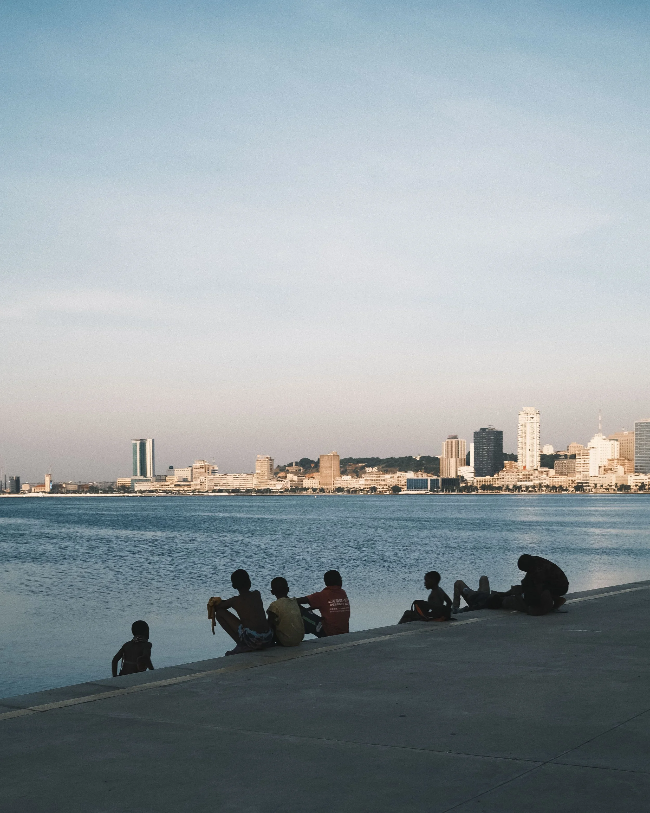 People sitting and relaxing near a body of water with a city skyline in the background during sunset or late afternoon.