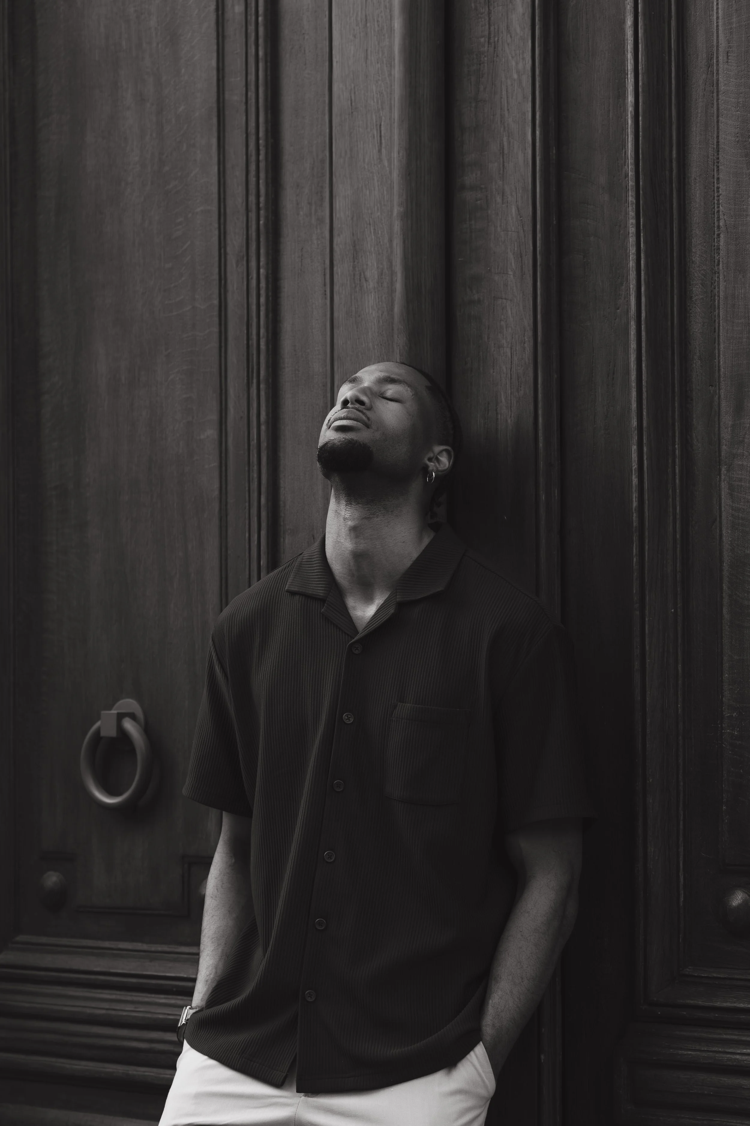 A man leaning against a dark wooden wall with closed eyes, wearing a dark short-sleeved shirt and light pants.