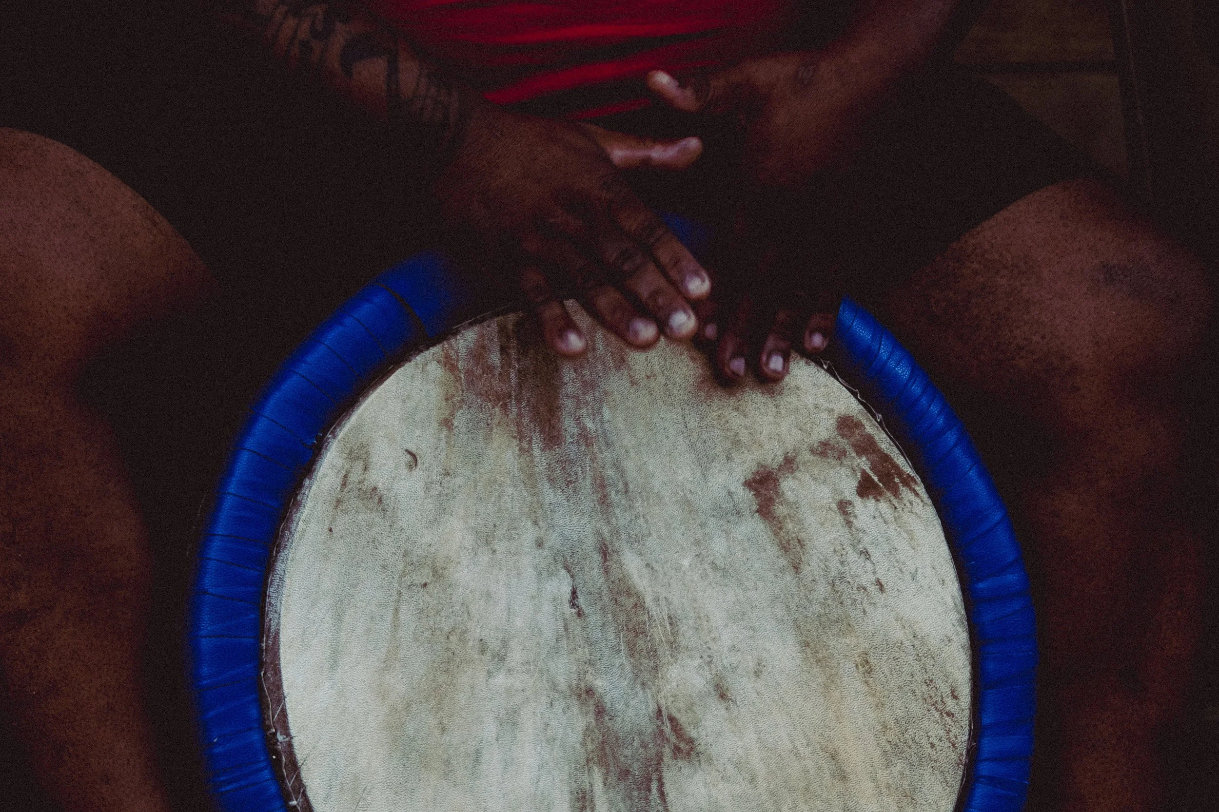 Two dark-skinned hands with tattoos clapping above a large drum with a worn white surface and a blue rim, seen from above.