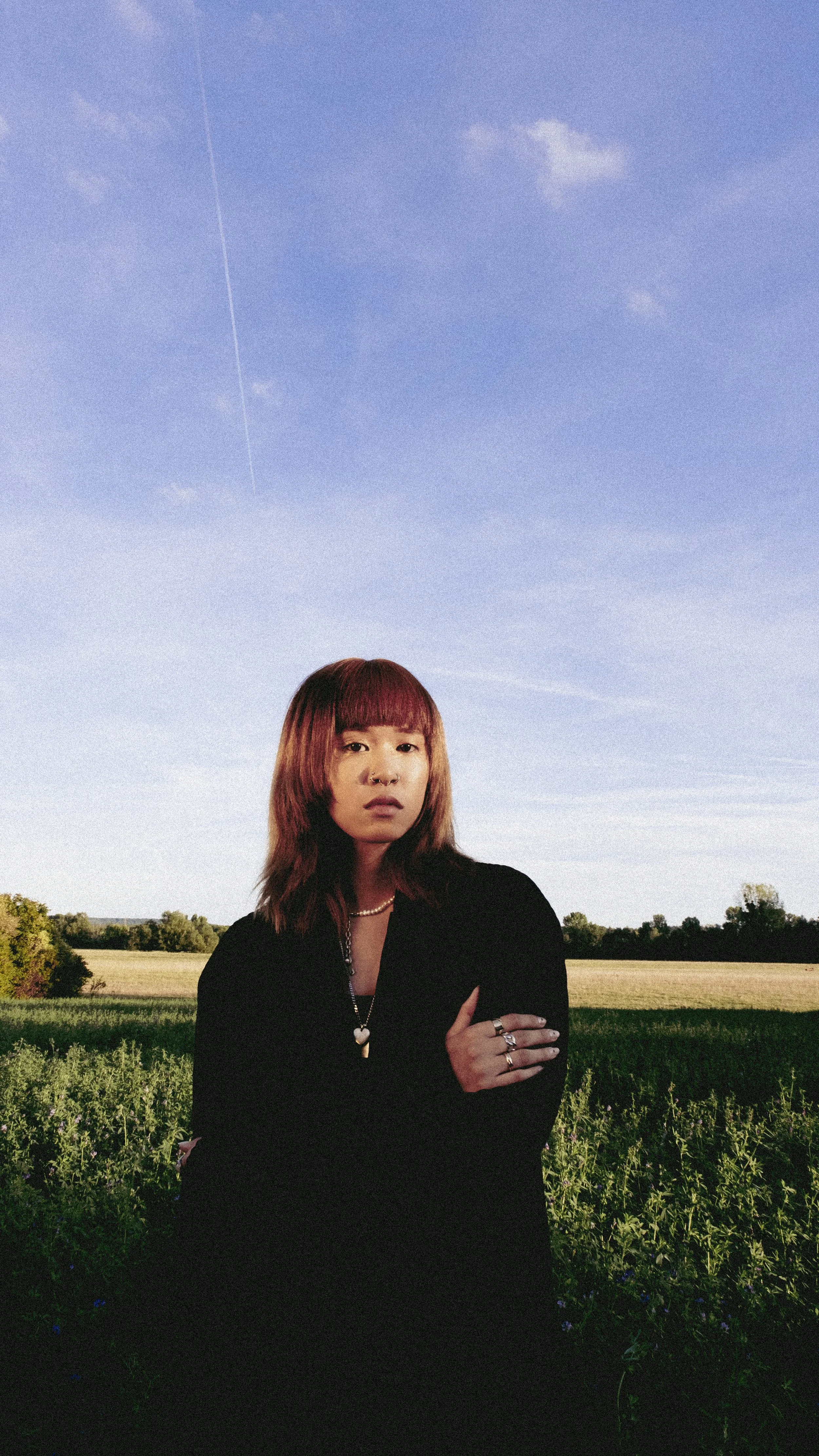 A woman with shoulder-length reddish-brown hair and bangs, wearing a black top and jewelry, standing outdoors in a field with a blue sky and wispy clouds above her.