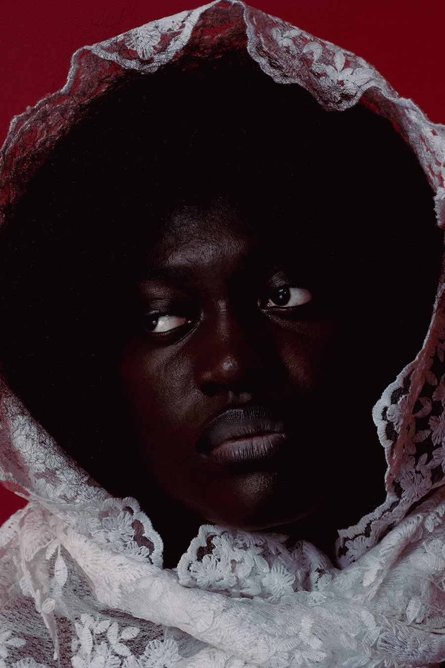 Close-up of a woman with dark skin, black hair, and piercing eyes, wearing a white lace hooded garment against a red background.
