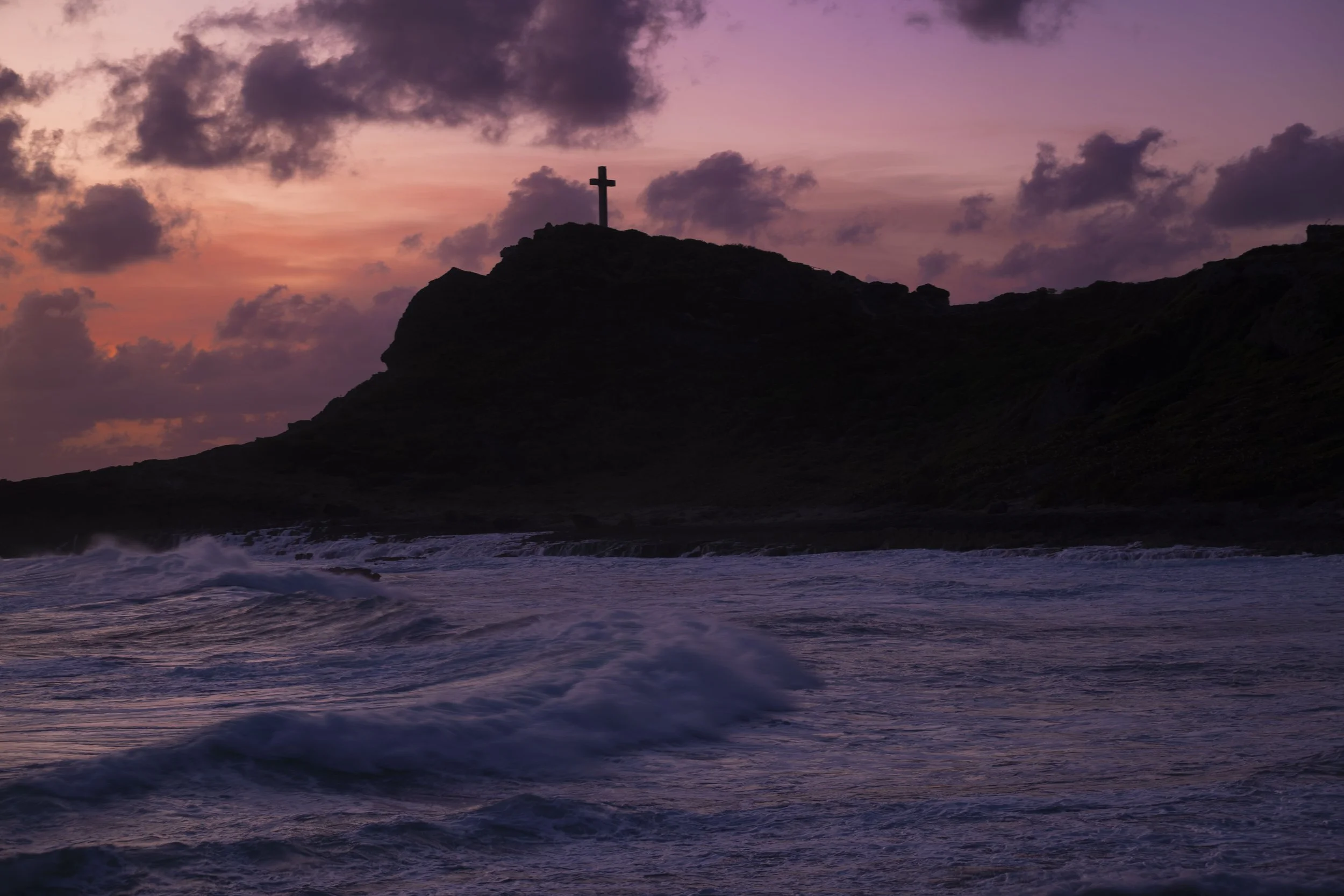 Silhouette of a hill with a cross on top, overlooking the ocean during sunset with purple and pink clouds