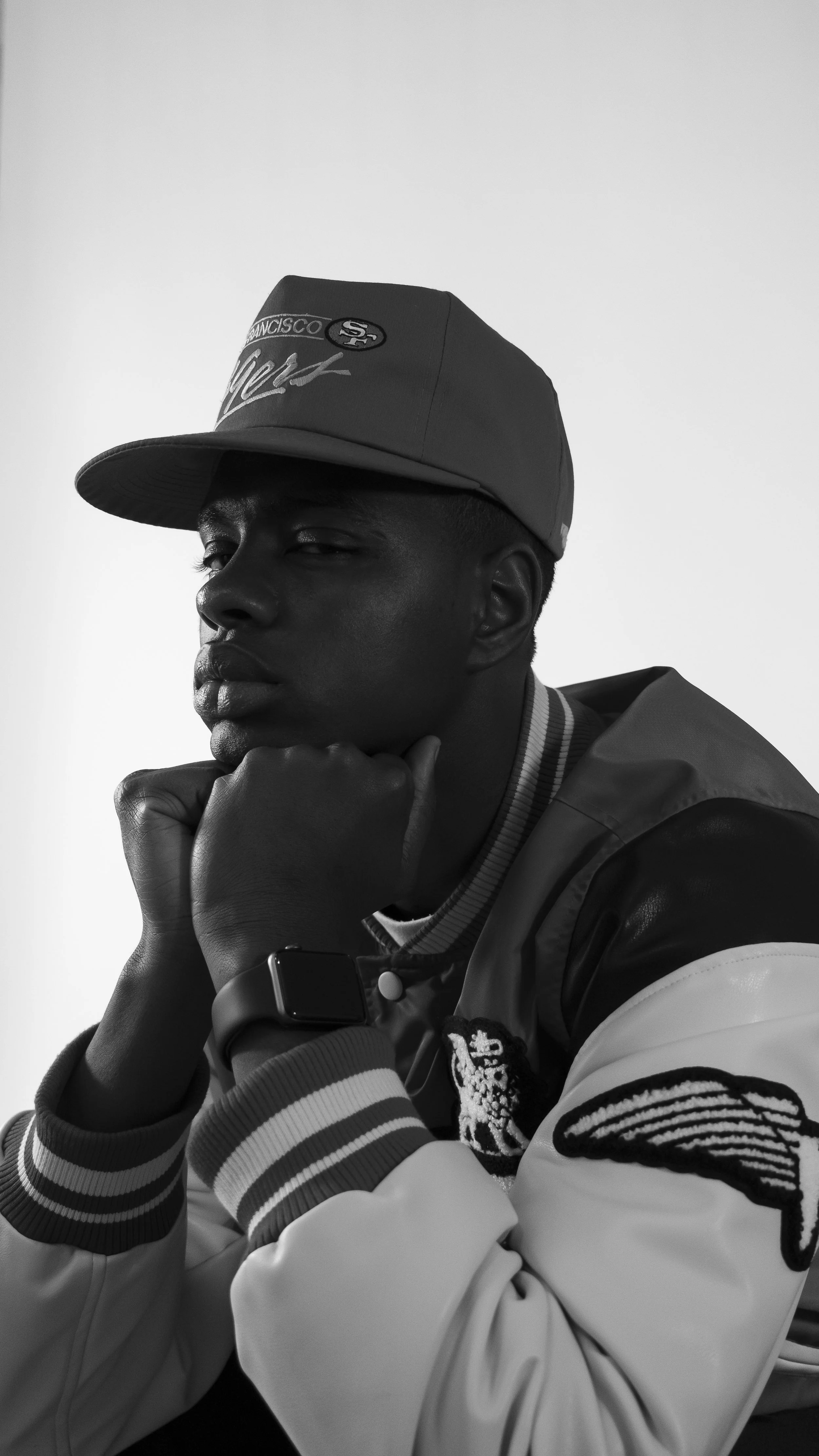 Black and white portrait of a young man wearing a cap, jacket, and smartwatch, resting his chin on his hand with a thoughtful expression.
