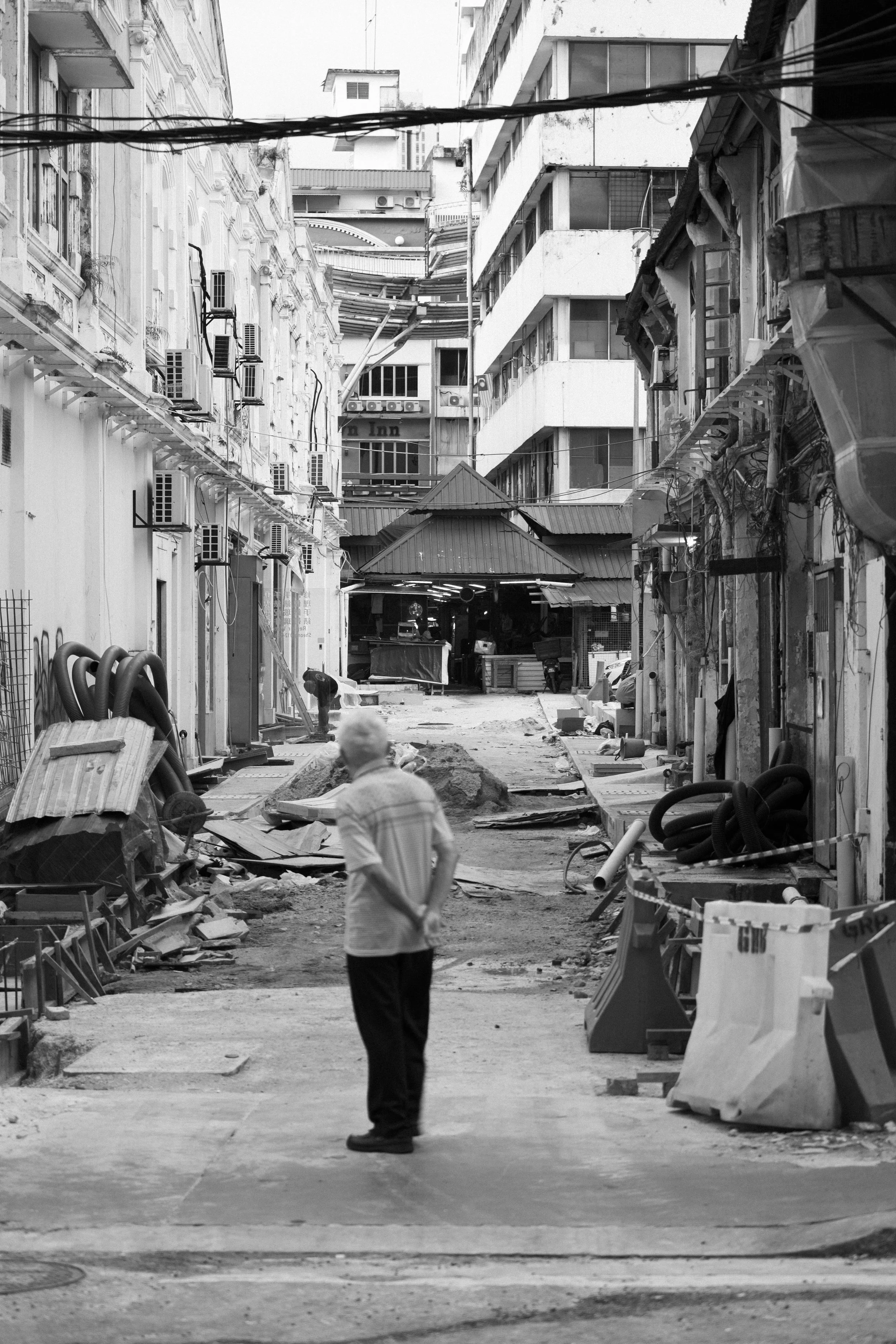 A man standing on a narrow urban street under construction, with debris, construction materials, and buildings on both sides.
