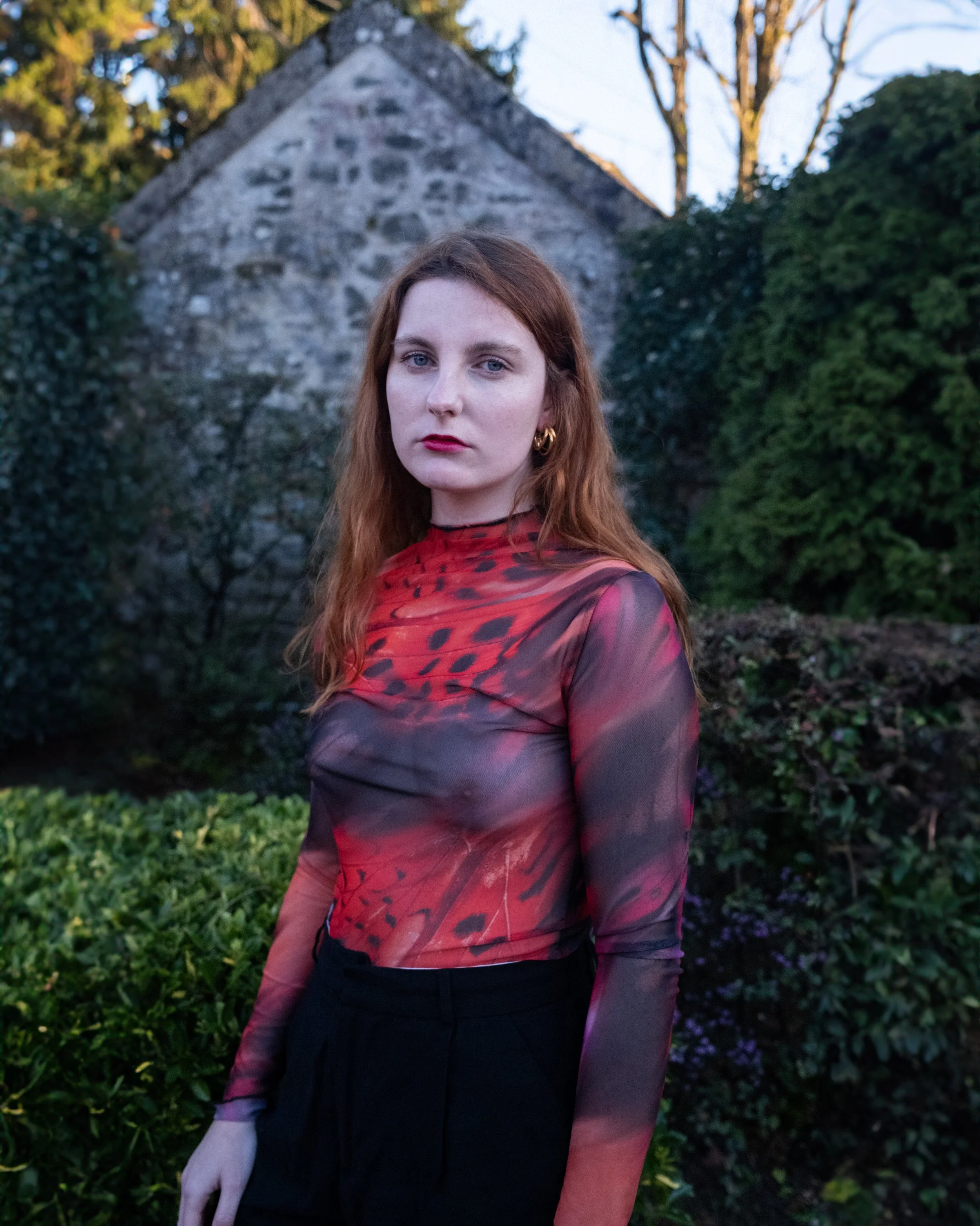 A woman with red hair and fair skin standing outdoors in front of a stone structure and green bushes, wearing a semi-transparent, long-sleeved red and black patterned top and black pants.