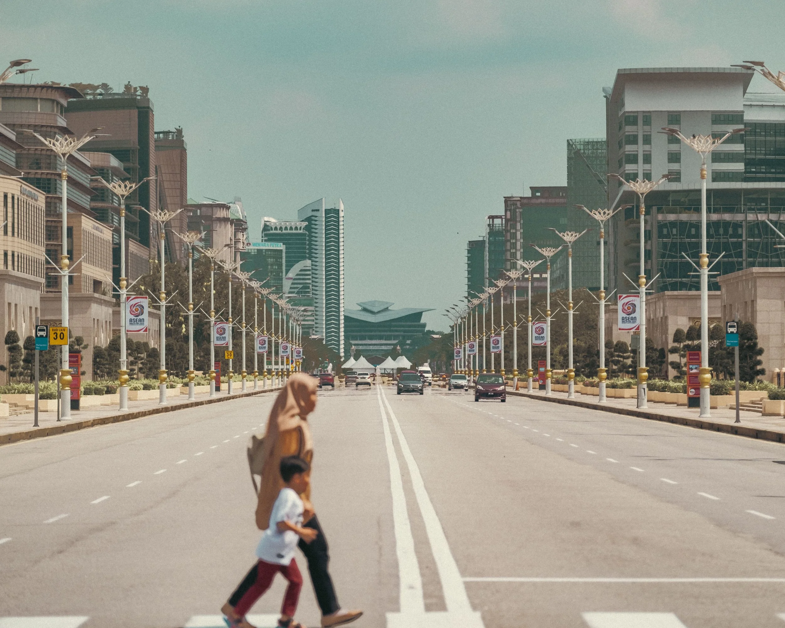 A woman wearing a hijab and a child crossing a wide, empty city street with tall buildings, street lamps, and flags on both sides, in the background.