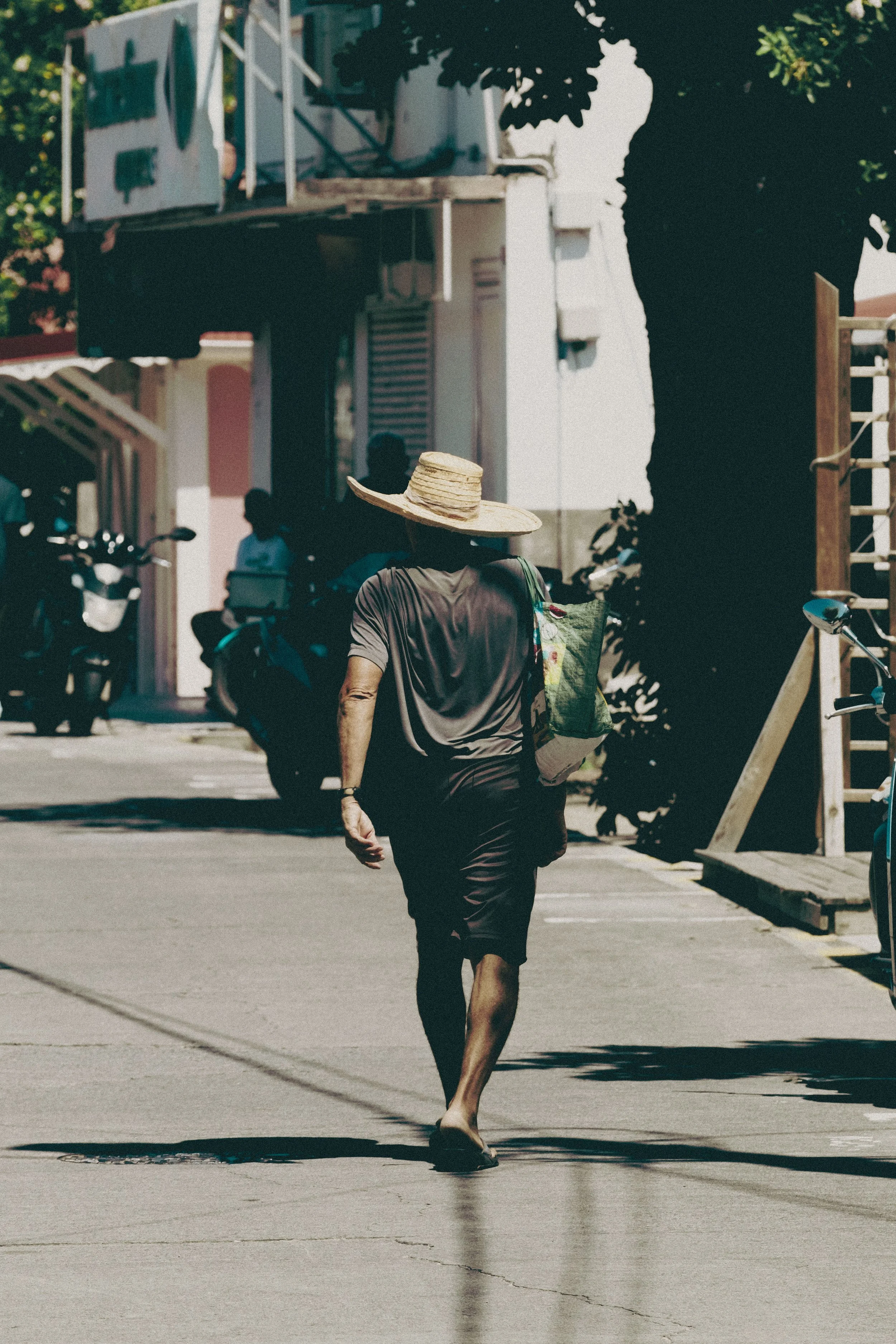 A person walking on a sidewalk carrying a large bag, wearing a wide-brimmed straw hat and dark clothing, with parked motorcycles and buildings in the background.