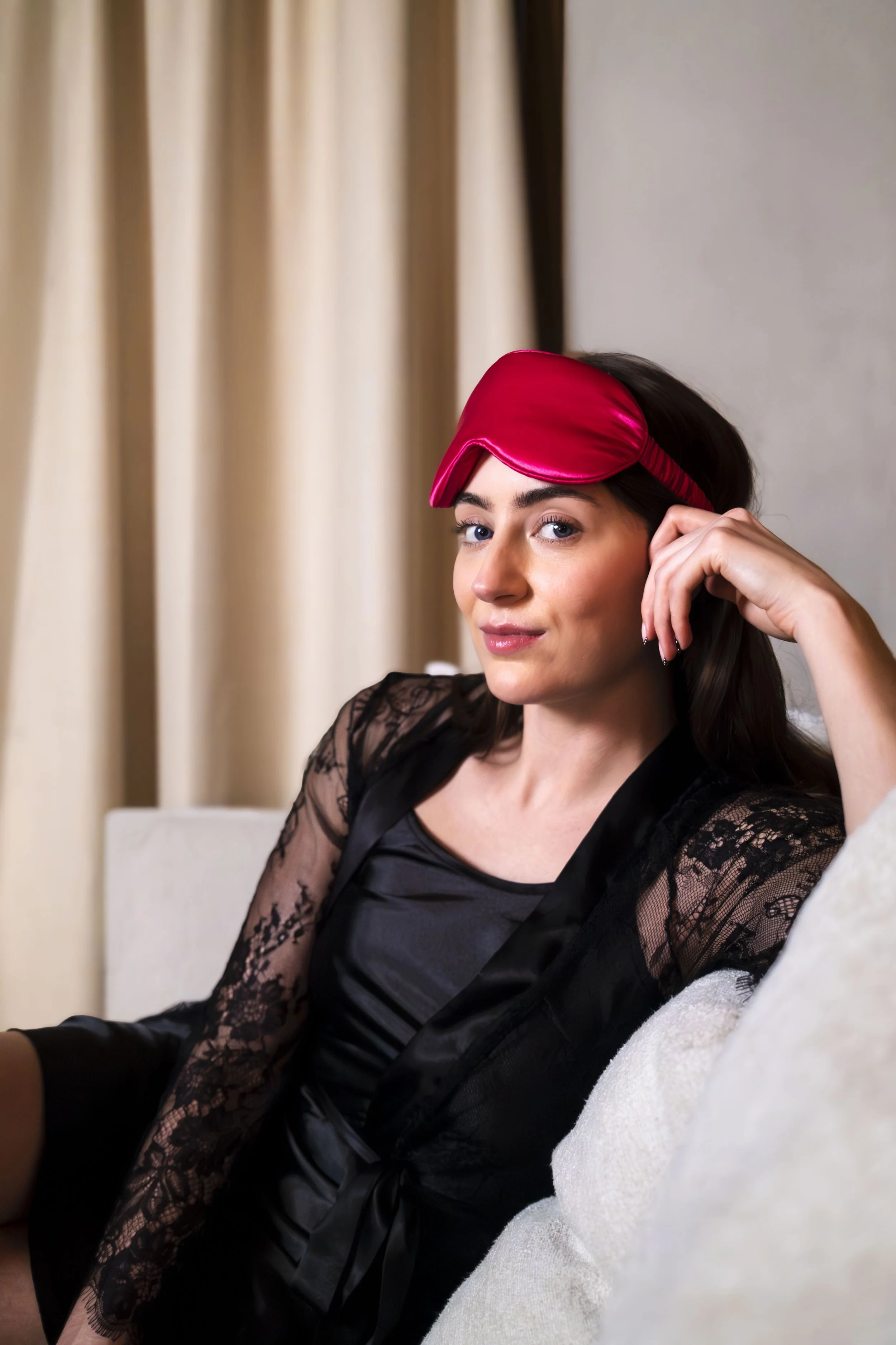 A woman wearing a red sleep mask on her forehead, sitting on a couch in a black lace dress, with beige curtains in the background.