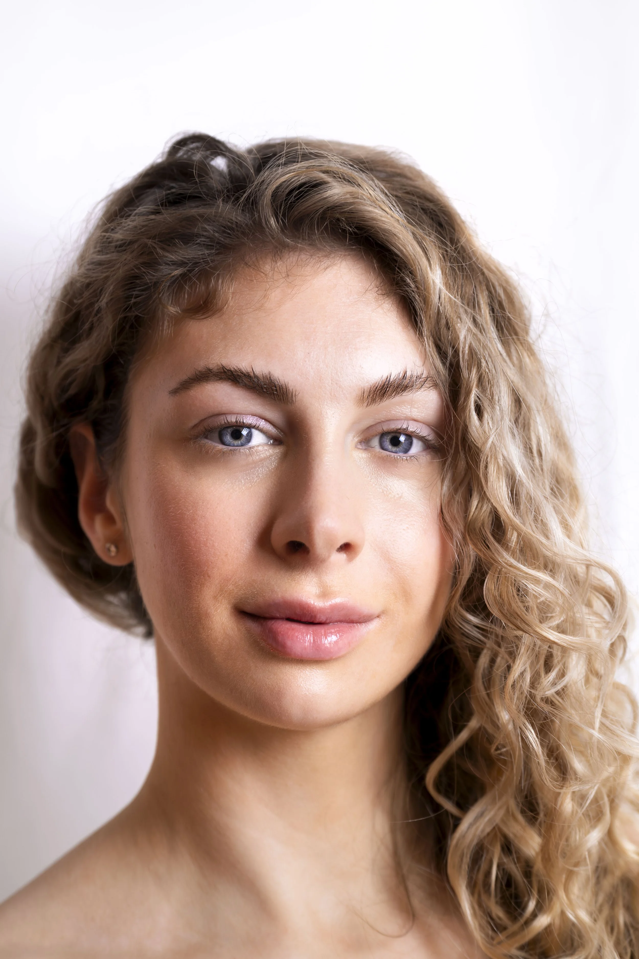 Close-up portrait of a young woman with curly blonde hair, fair skin, and blue eyes, smiling softly against a plain white background.