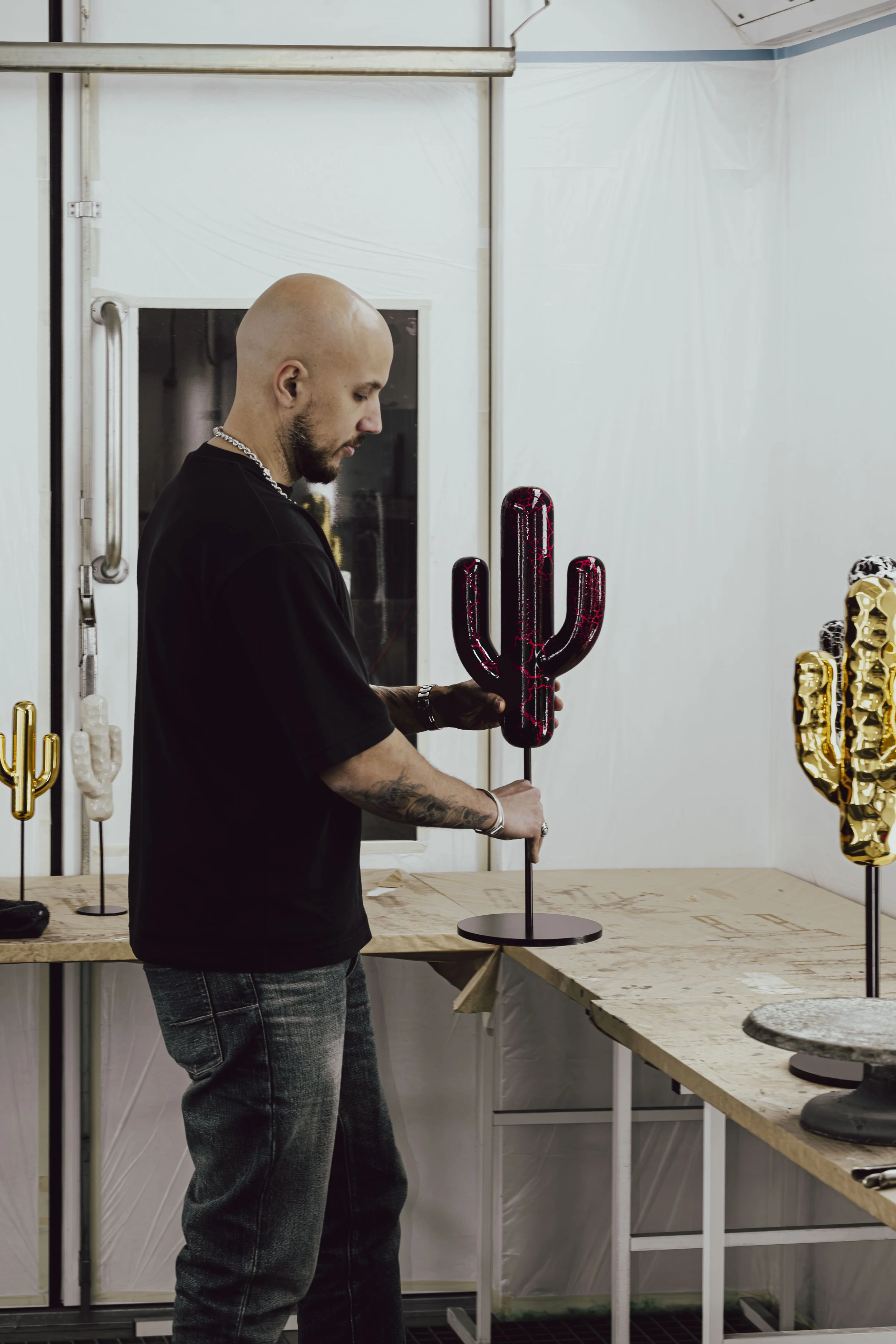 Man with bald head and tattoos holding a black and red cactus-shaped sculpture in an art studio.