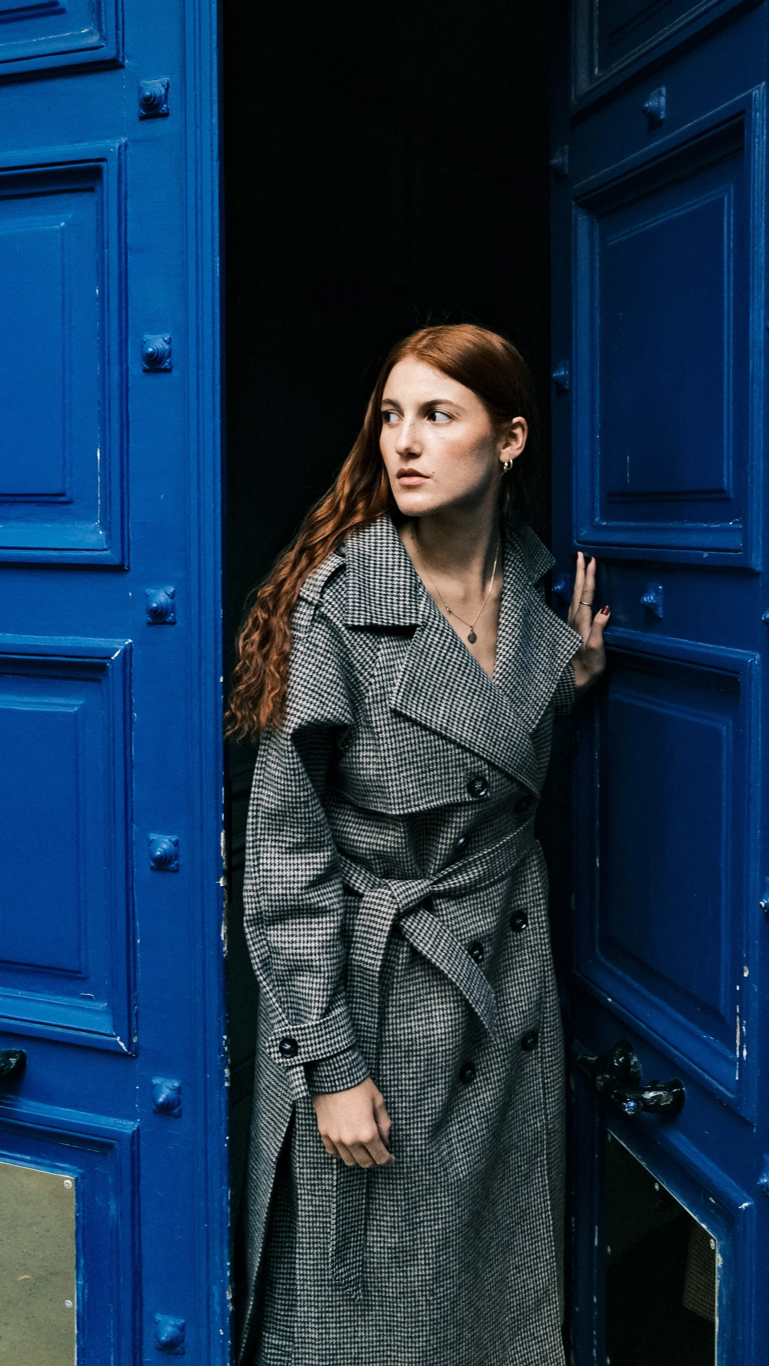 A young woman with long red hair peeking out from a doorway flanked by bright blue walls.
