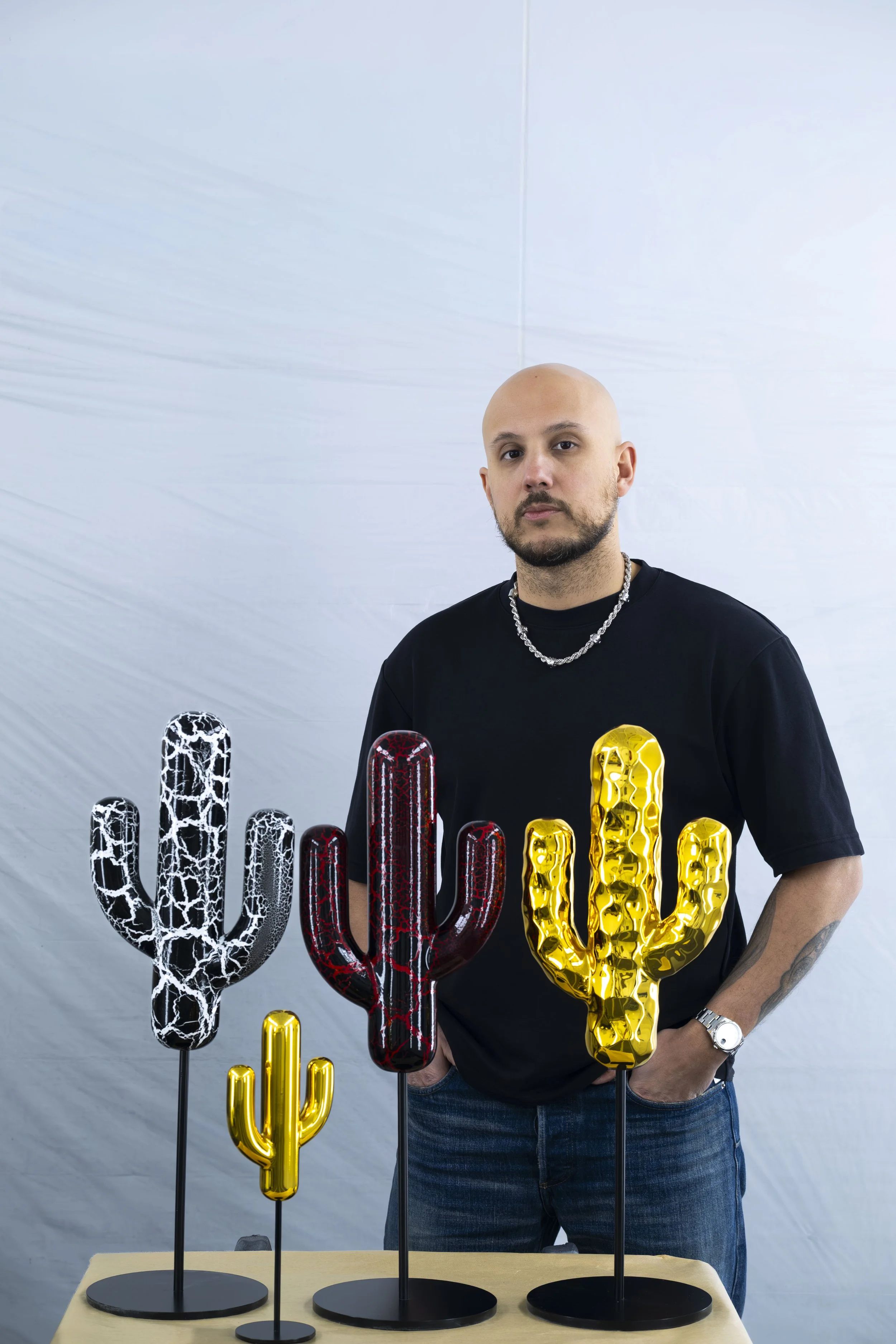 A man with a shaved head, beard, wearing a black T-shirt, silver chain, and watch, standing behind a table with four decorative cactus sculptures in black, red, and yellow colors.