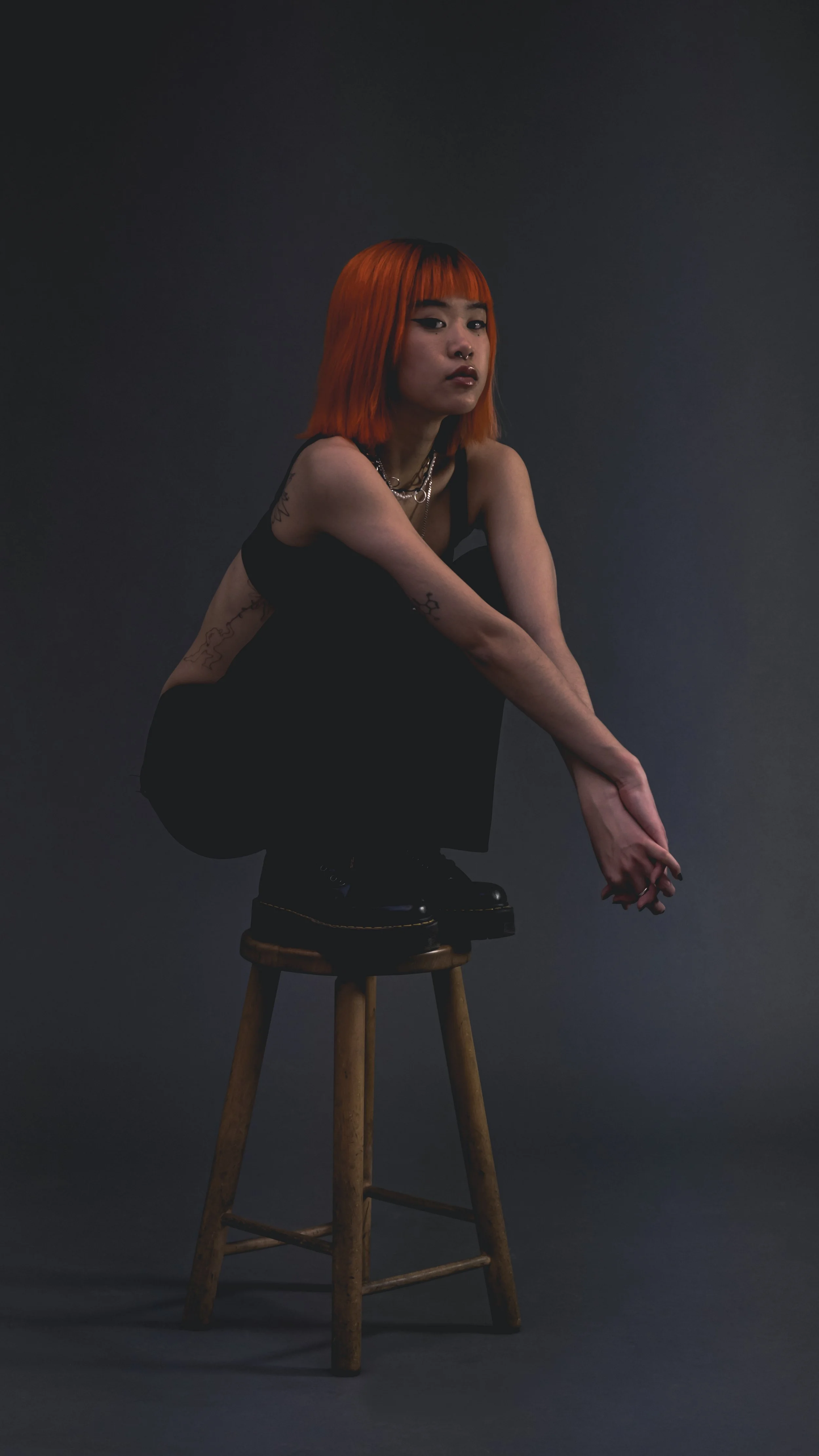 A young woman with orange hair, tattoos, and wearing black clothing, crouches on a wooden stool against a dark, solid background.