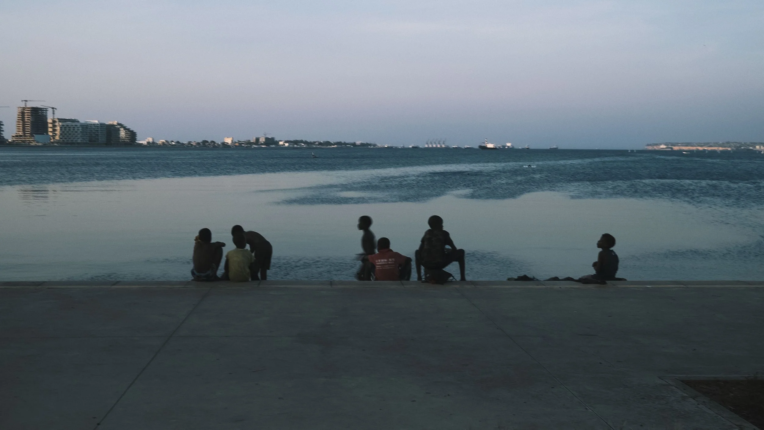Six children sitting and standing by the water's edge in a coastal city at dusk, with buildings and ships visible across the bay.