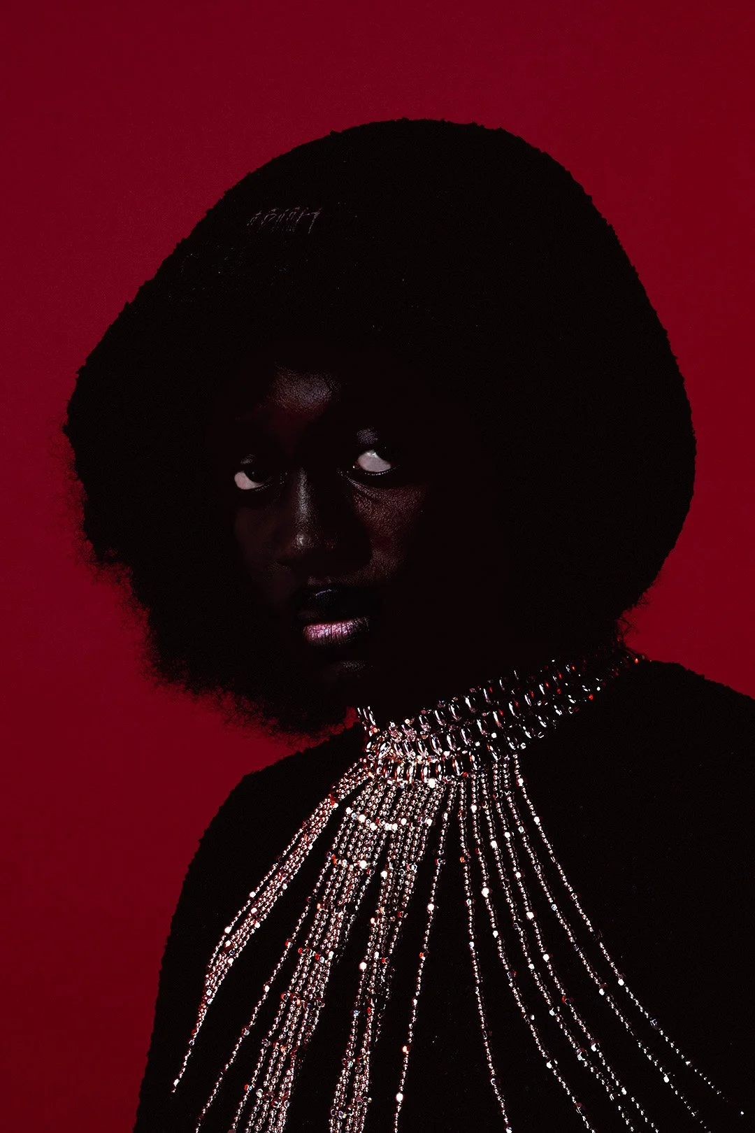 Portrait of a woman with dark skin wearing a large black afro hairstyle, adorned with a shiny, multi-layered silver chain necklace, against a solid red background.