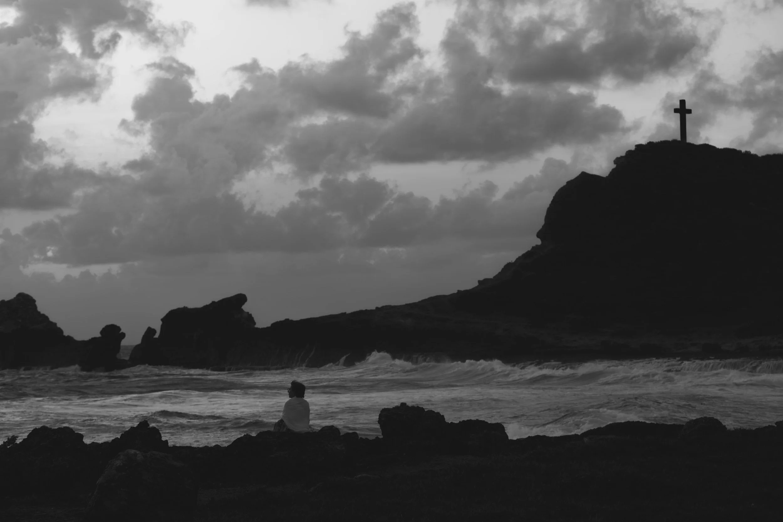 A person sitting on rocks by the ocean, with a large hill topped by a cross in the background, under a cloudy sky.