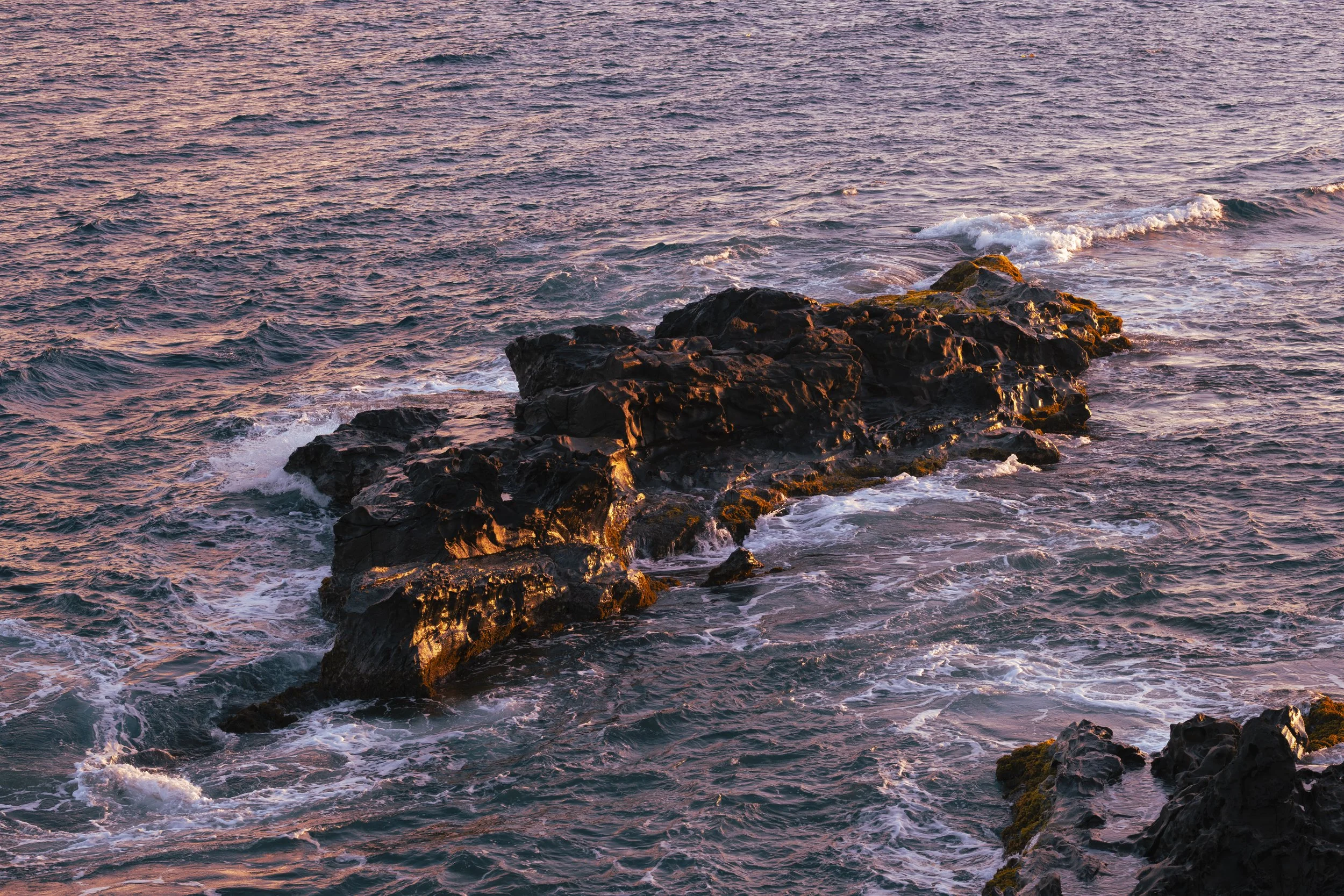 Rocks in the ocean with waves crashing around them during sunset or sunrise, casting a warm glow.
