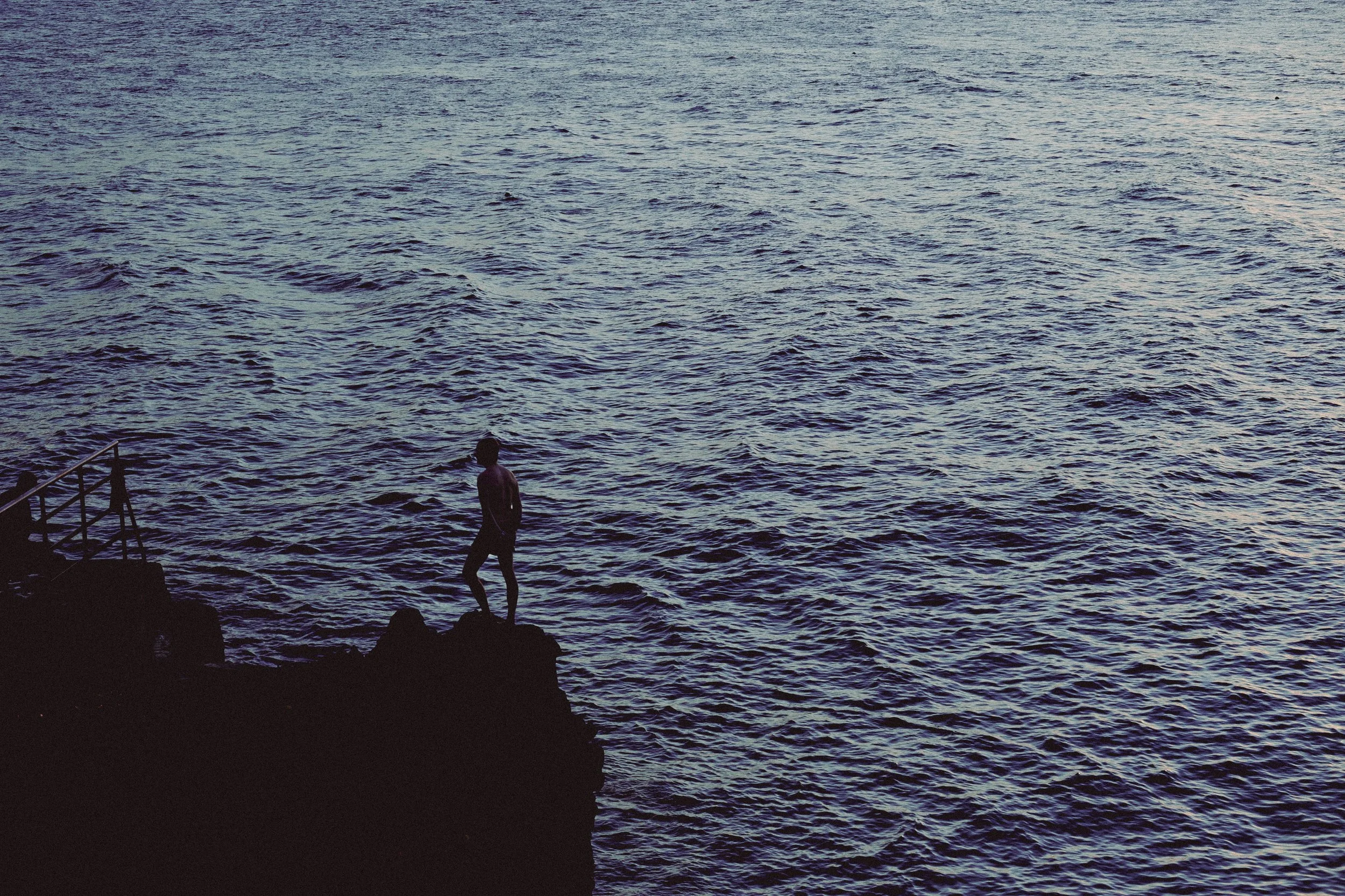 A person standing on rocks by the water during sunset or dusk, with the ocean stretching out into the horizon.
