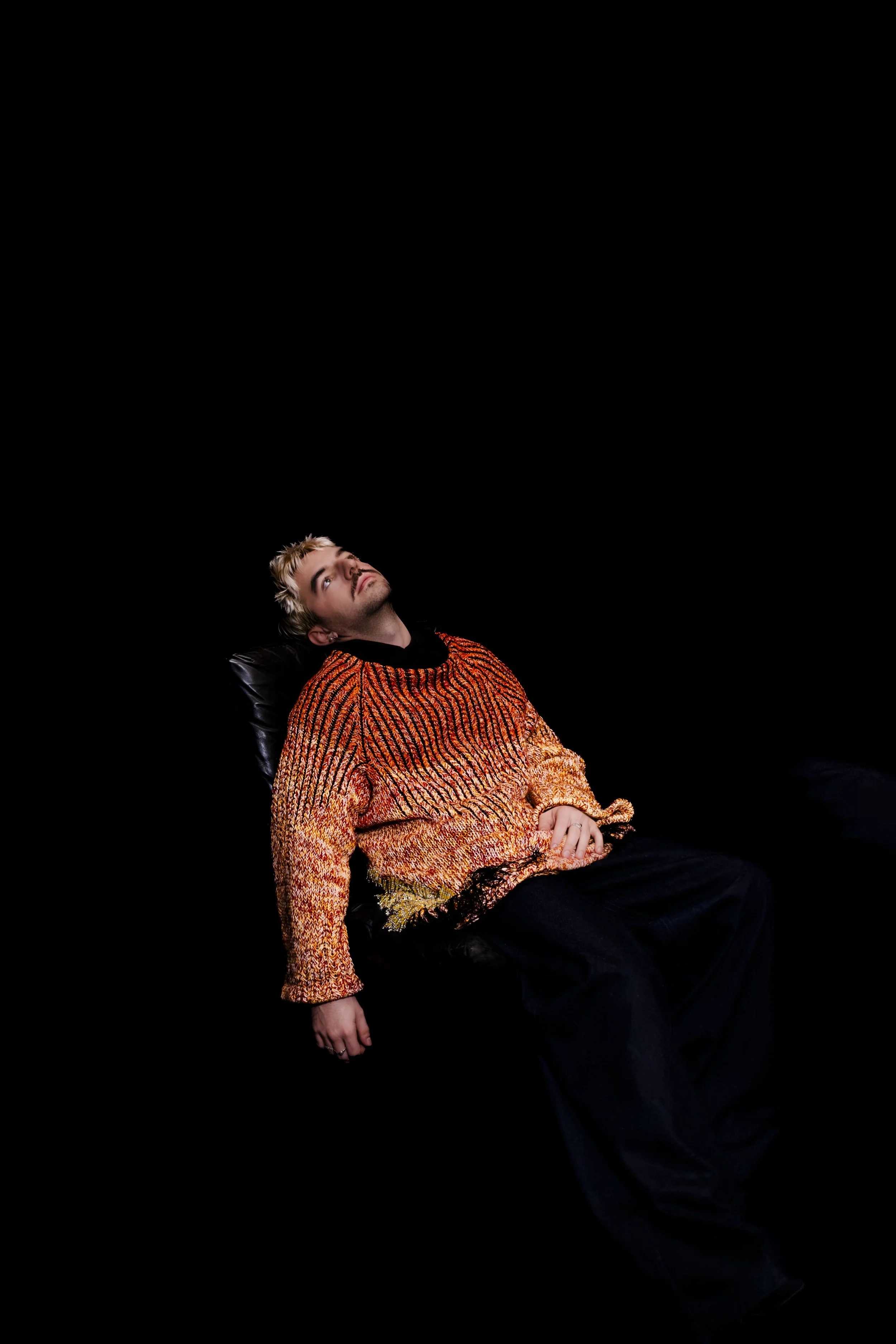 A young man with blond hair and a beard, wearing a multicolored knit sweater, is sitting in a black chair against a black background. He is leaning back and looking upwards.