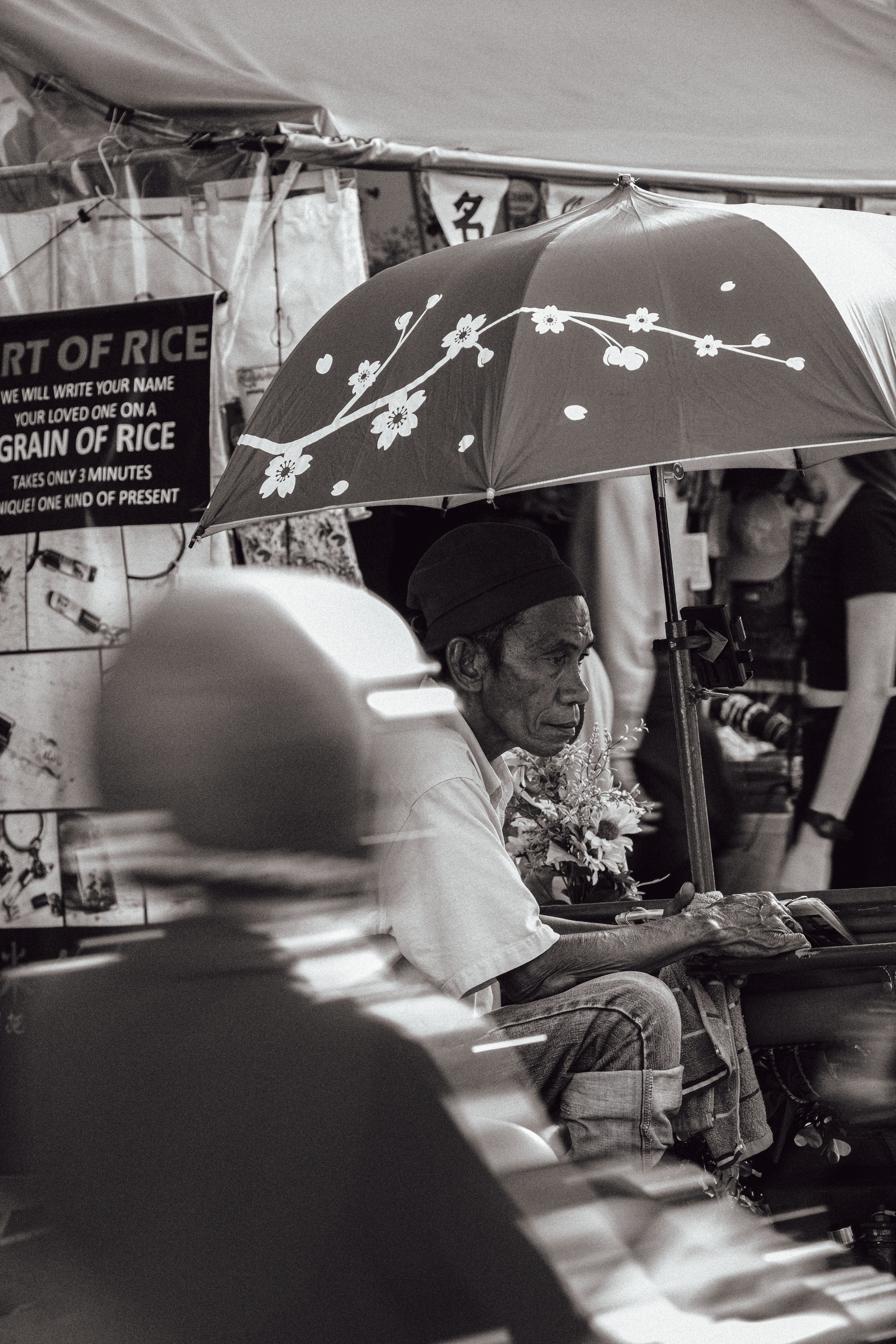 An elderly man sitting under a floral umbrella at a market stall, surrounded by signs and other shoppers.