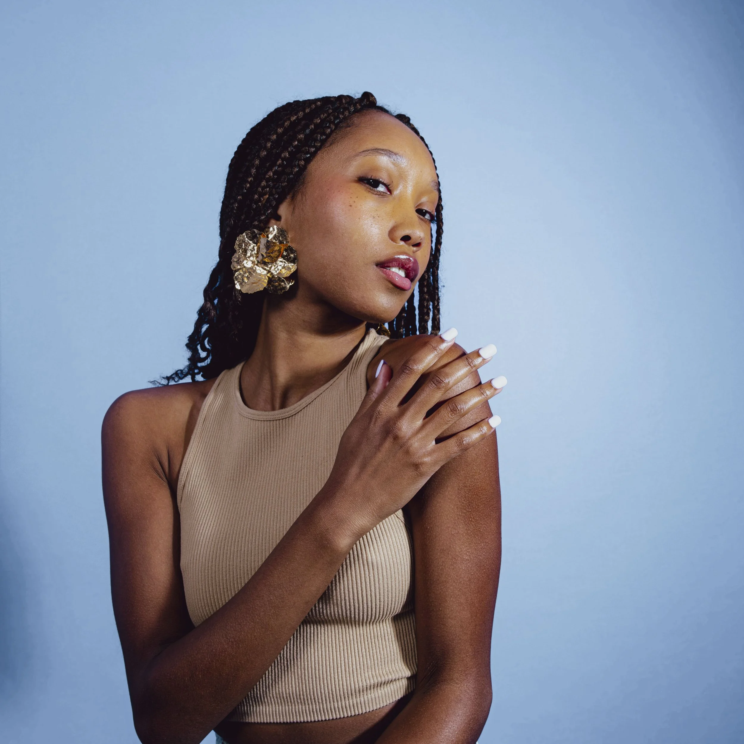 A woman with braided hair and large gold earrings poses against a blue background, wearing a beige sleeveless top and white nail polish.