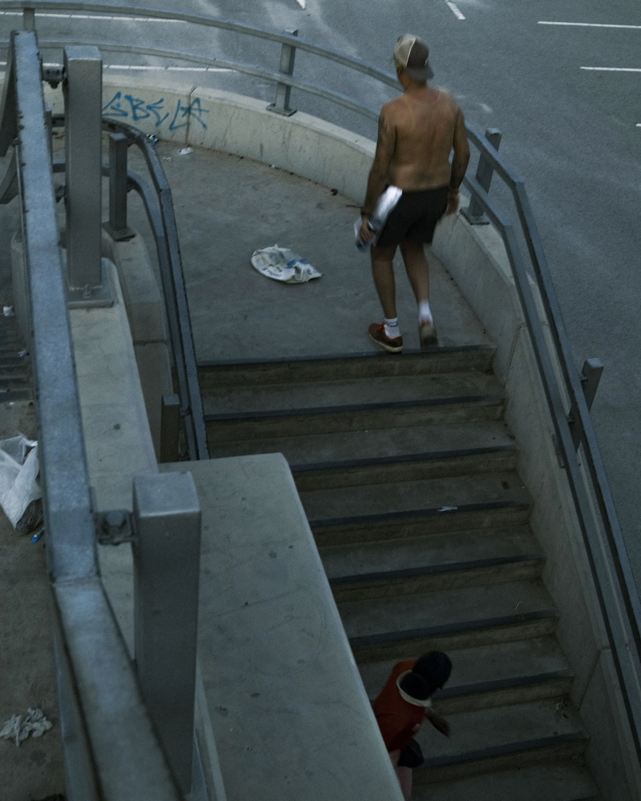 A shirtless man wearing shorts, a hat, and sneakers walking down stairs near a concrete sidewalk with graffiti, and a person sitting on the stairs wearing a red hoodie.