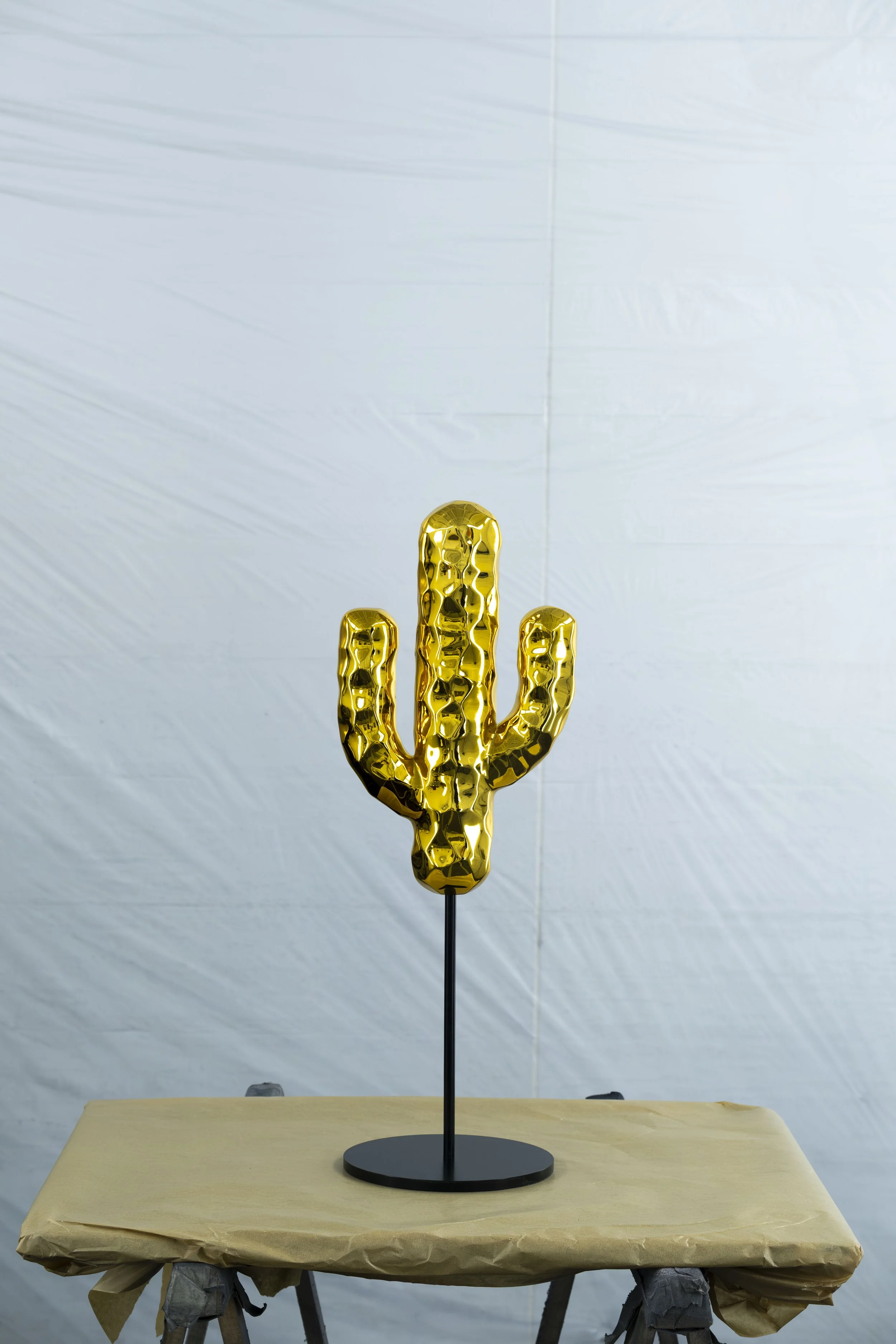 A decorative sculpture of a cactus made of shiny, gold-colored metal on a black stand, placed on a table covered with brown paper.