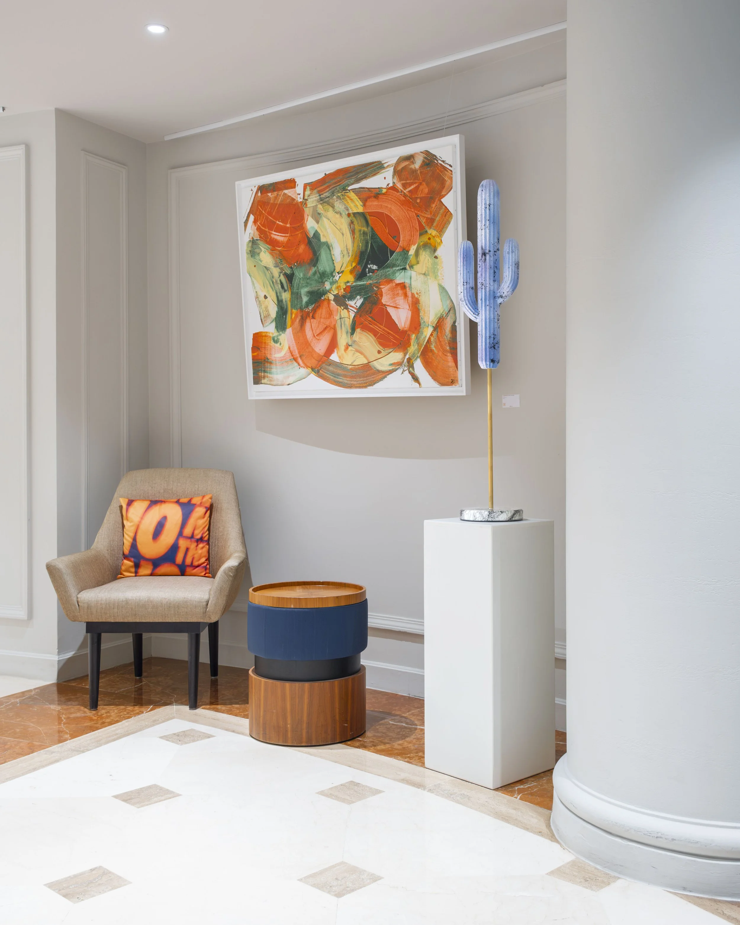 Interior of an art gallery featuring a colorful abstract painting on the wall, a beige chair with a vibrant pillow, a small round table with a two-tone wooden and blue design, and a modern blue cactus sculpture on a white pedestal.