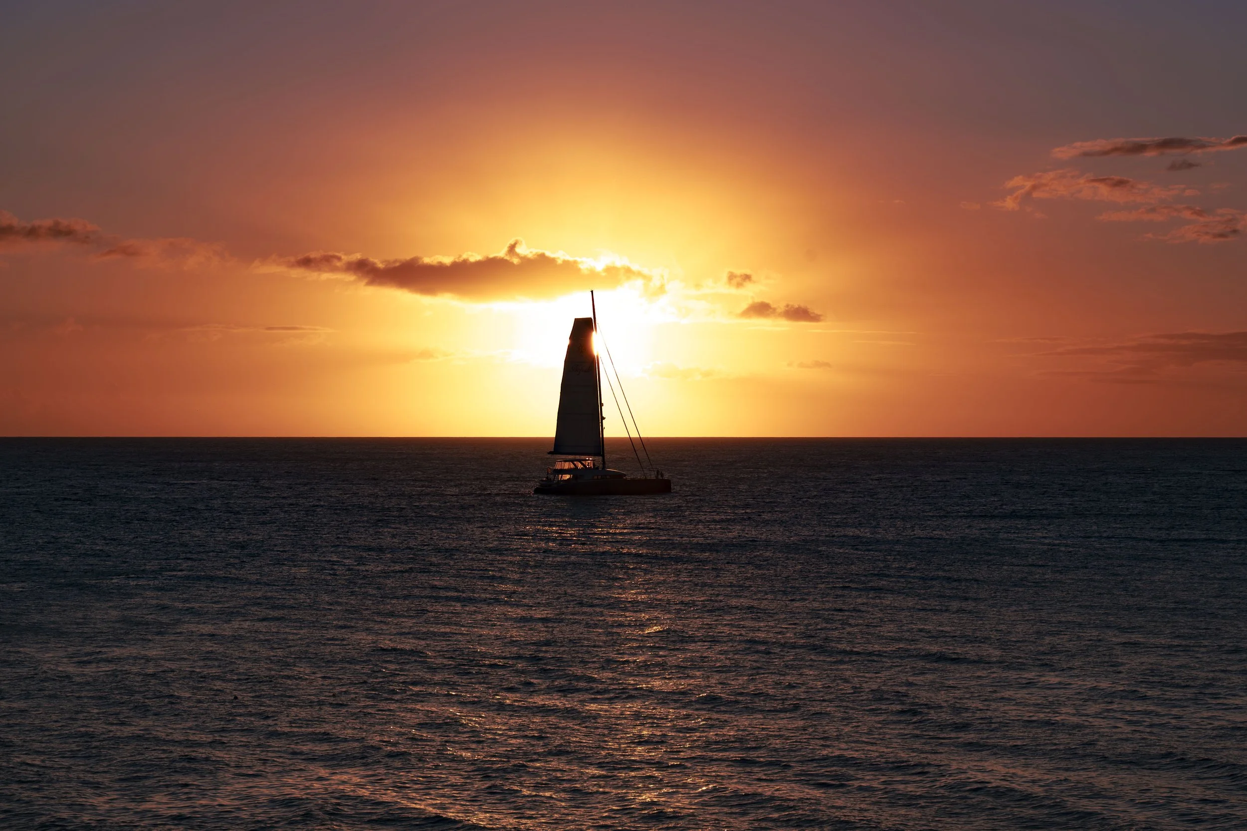 A sailboat on the ocean during a vibrant sunset with orange, pink, and purple hues in the sky, and a few scattered clouds.