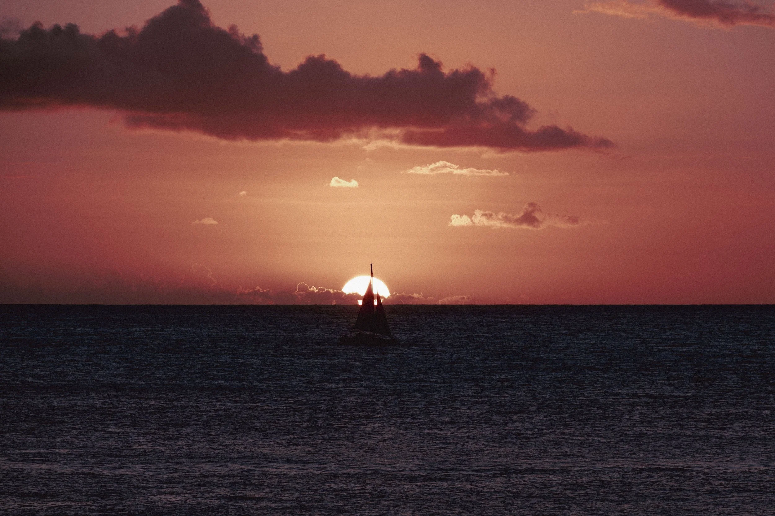 A sailboat sailing in the ocean during sunset with the sun partially below the horizon, pink and orange sky, and dark clouds.