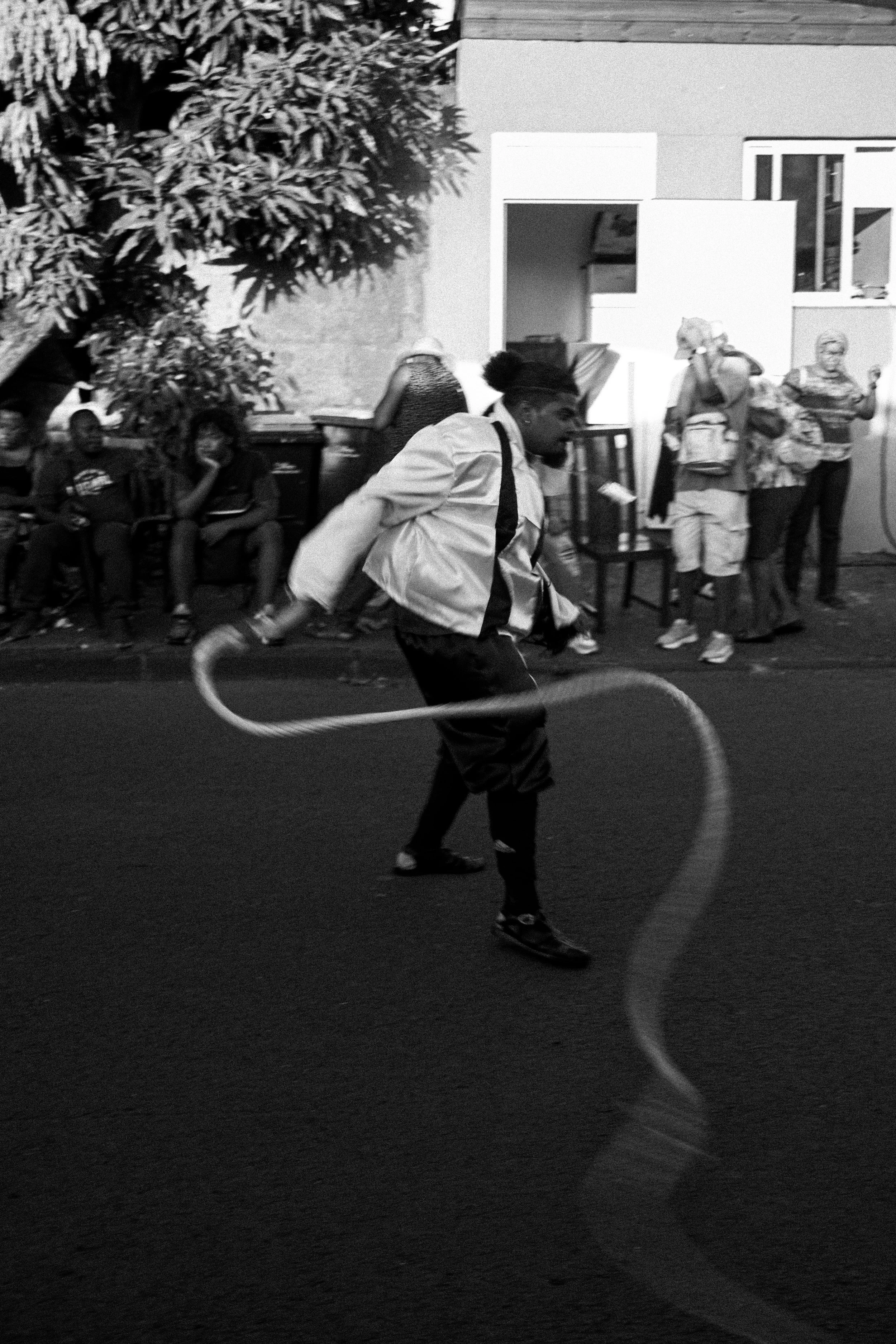 A young person jumping rope on a city street at night, with several spectators sitting and standing in the background.
