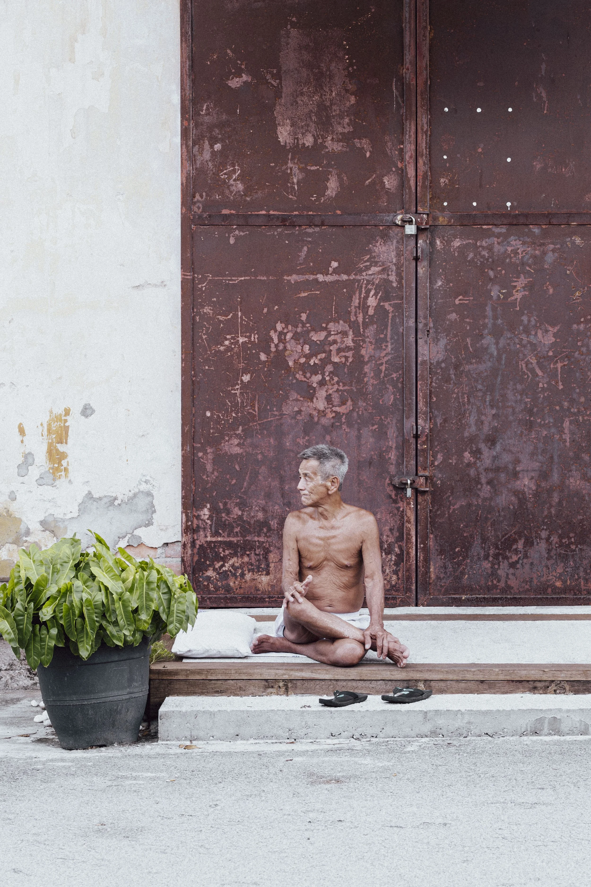 An elderly shirtless man sitting cross-legged on a concrete step in front of a large, rusty metal door. A white pillow and black sandals are beside him, and there is a large green potted plant to his left. The wall behind him is partially painted.
