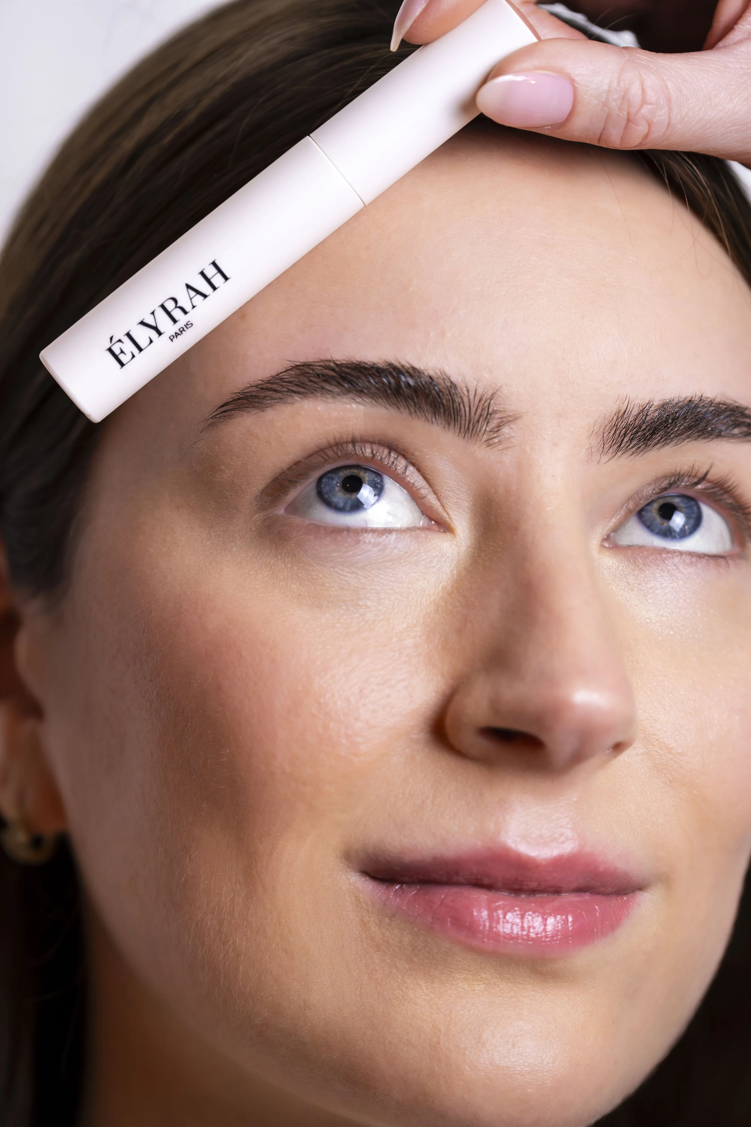 Close-up of a woman with fair skin and blue eyes applying a white skincare or cosmetic product labeled 'ÉLYRAH PARIS' to her forehead with her fingers.