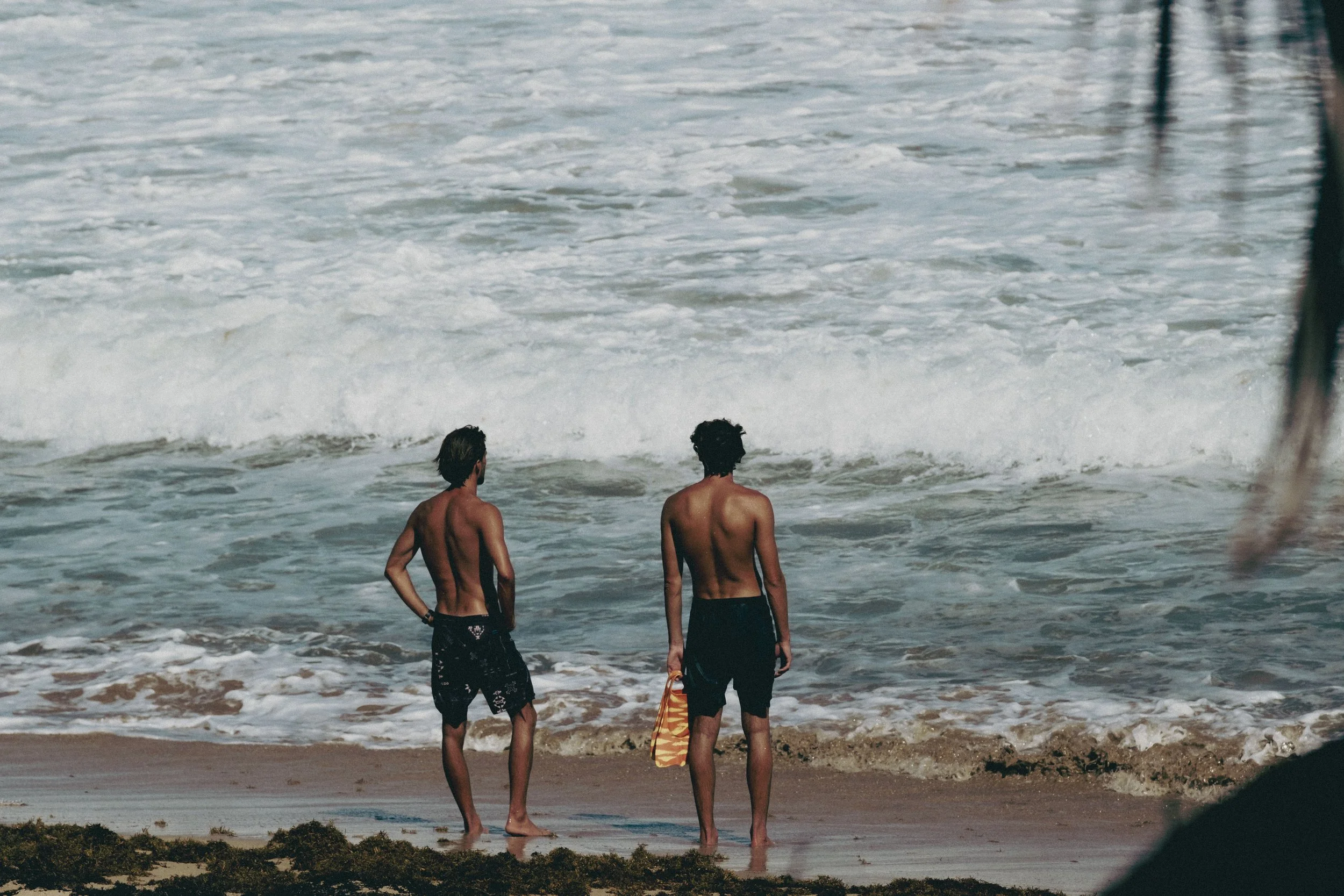 Two shirtless young men with dark hair standing on a sandy beach near the ocean, one holding a colorful towel, facing the waves.
