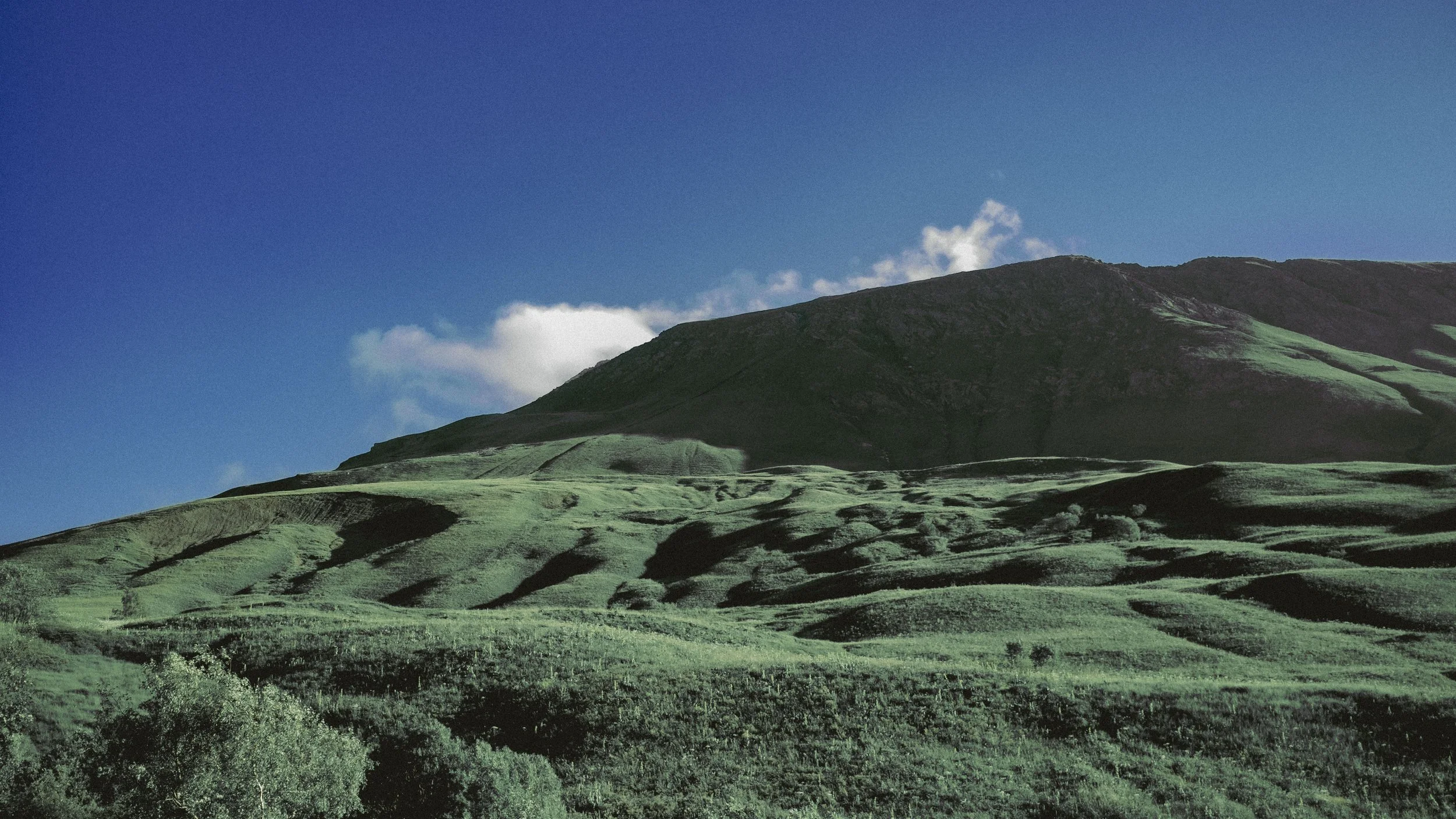 Green rolling hills and a mountain under a blue sky with some clouds.