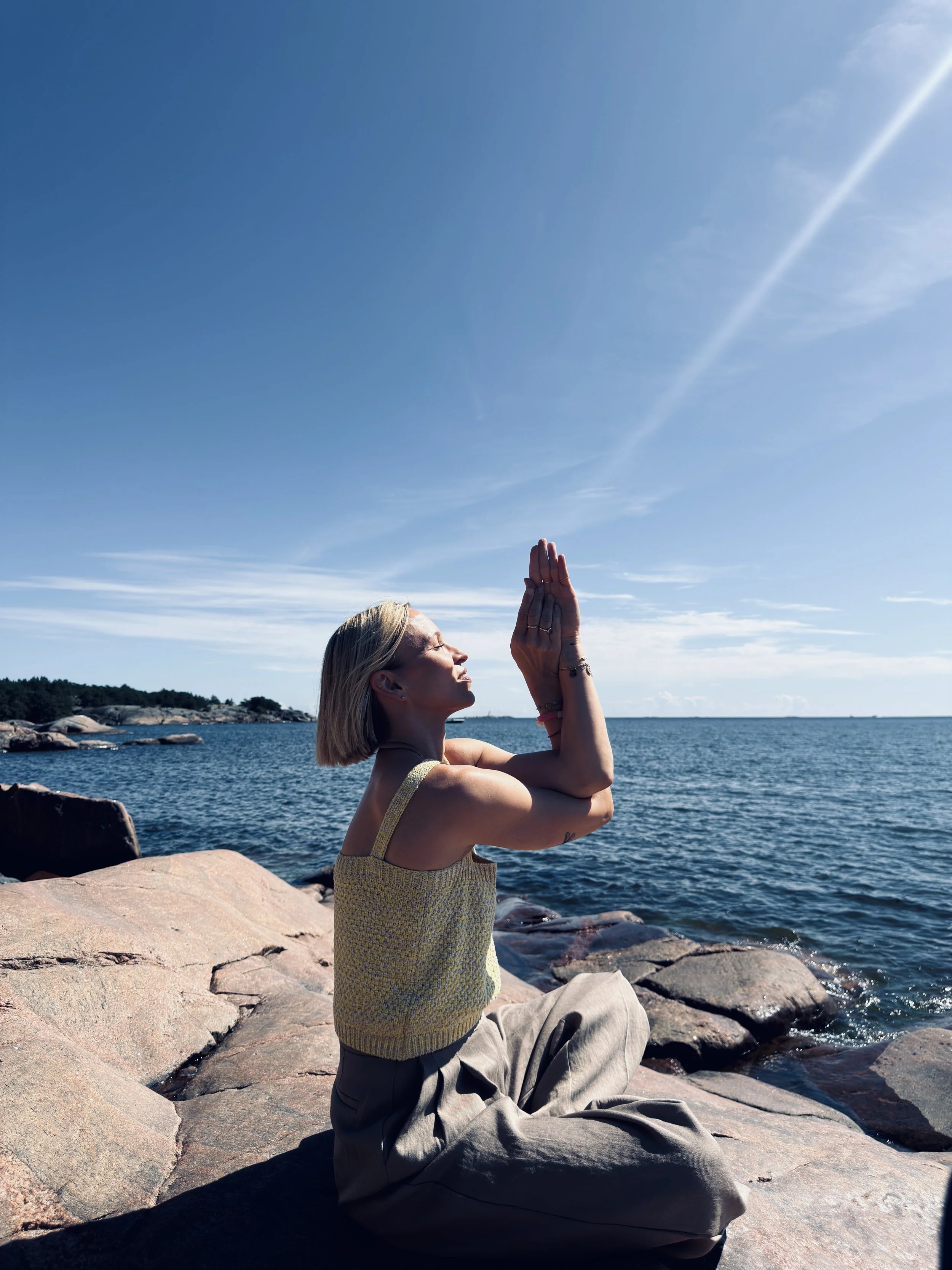 Nainen istuu kallioilla meren rannalla aurinkoisella säällä, tekee joogaliikkeitä ja näyttää rauhalliselta.