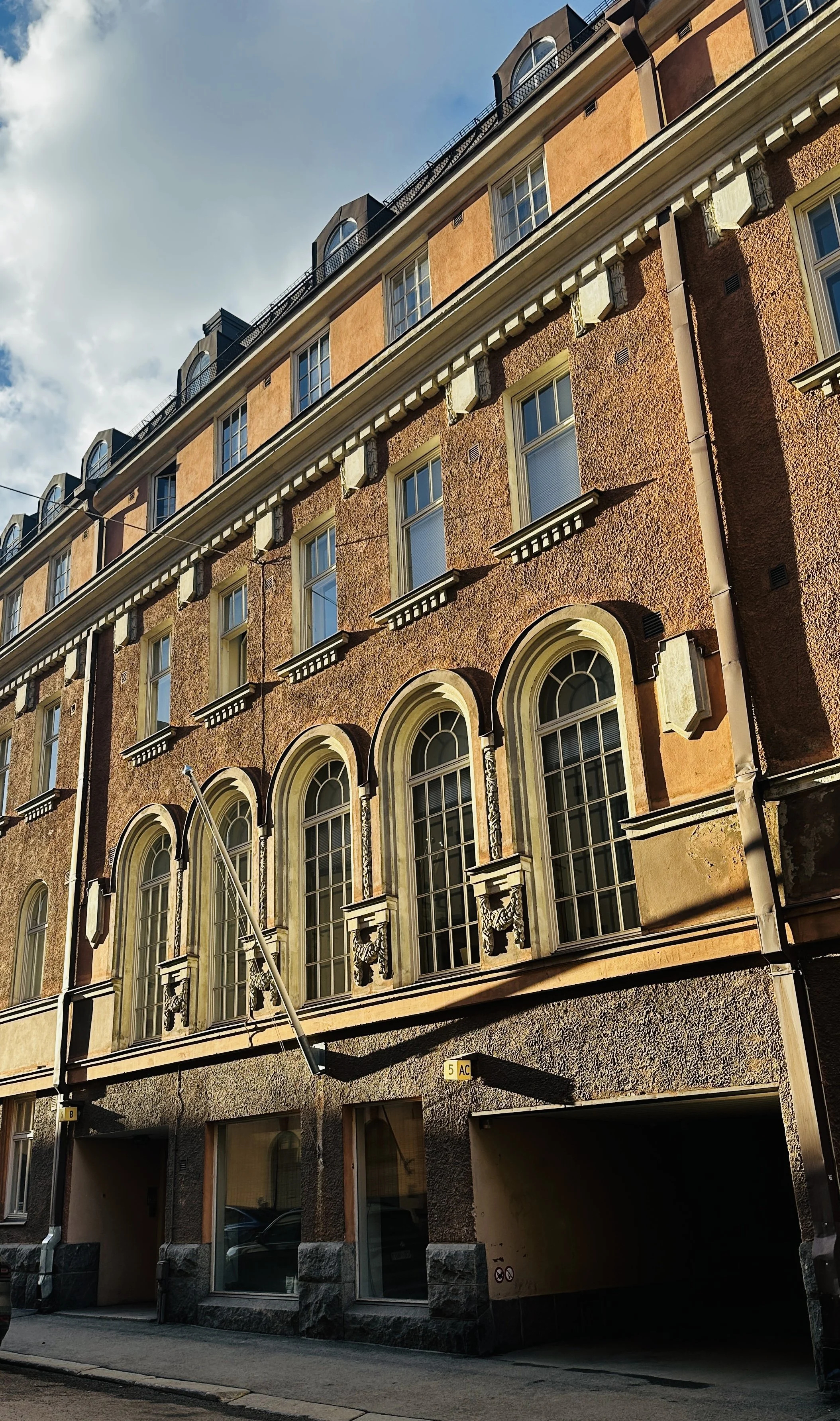A tall, historic multi-story brick building with large arched windows on the lower floors and rectangular windows on the upper floors, ornate architectural details, and a rough textured facade. The sky is partly cloudy and the photo is taken from the street level.