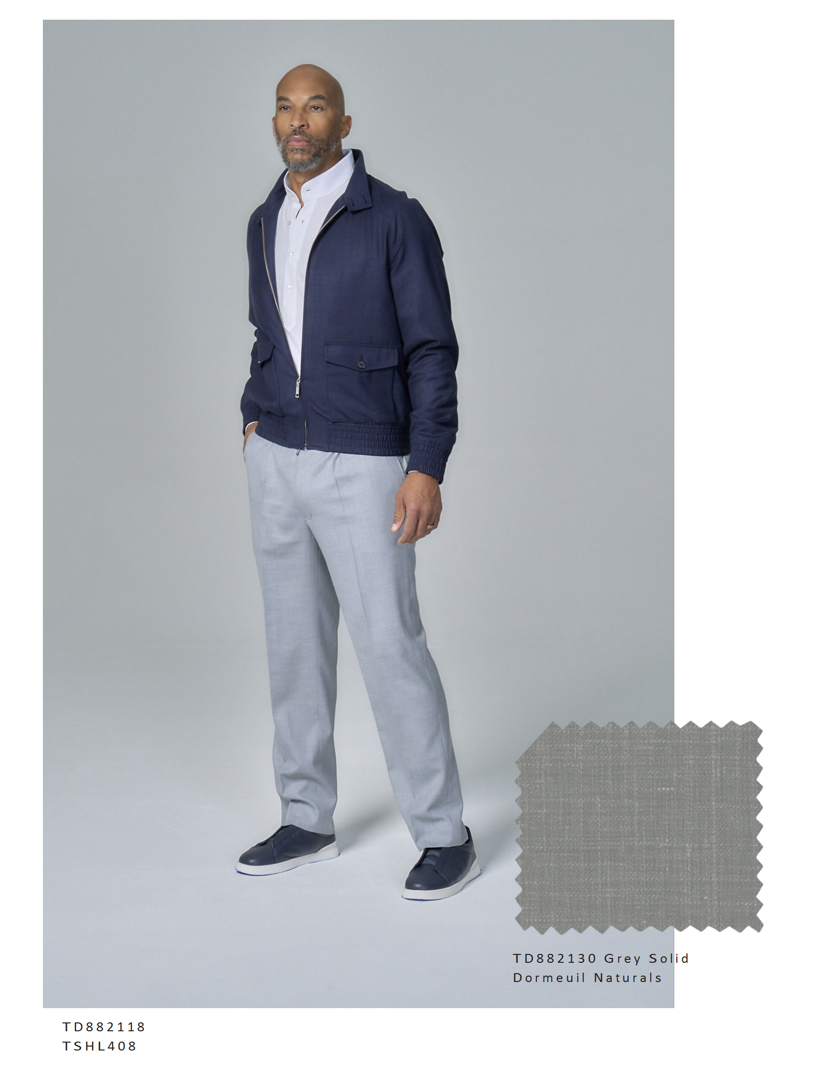 A man standing against a plain light gray background, dressed in a navy blue zip-up jacket, light gray trousers, and navy slip-on shoes. There is a fabric swatch labeled 'TD882130 Grey Solid Dormeuil Naturals' in the lower right corner.