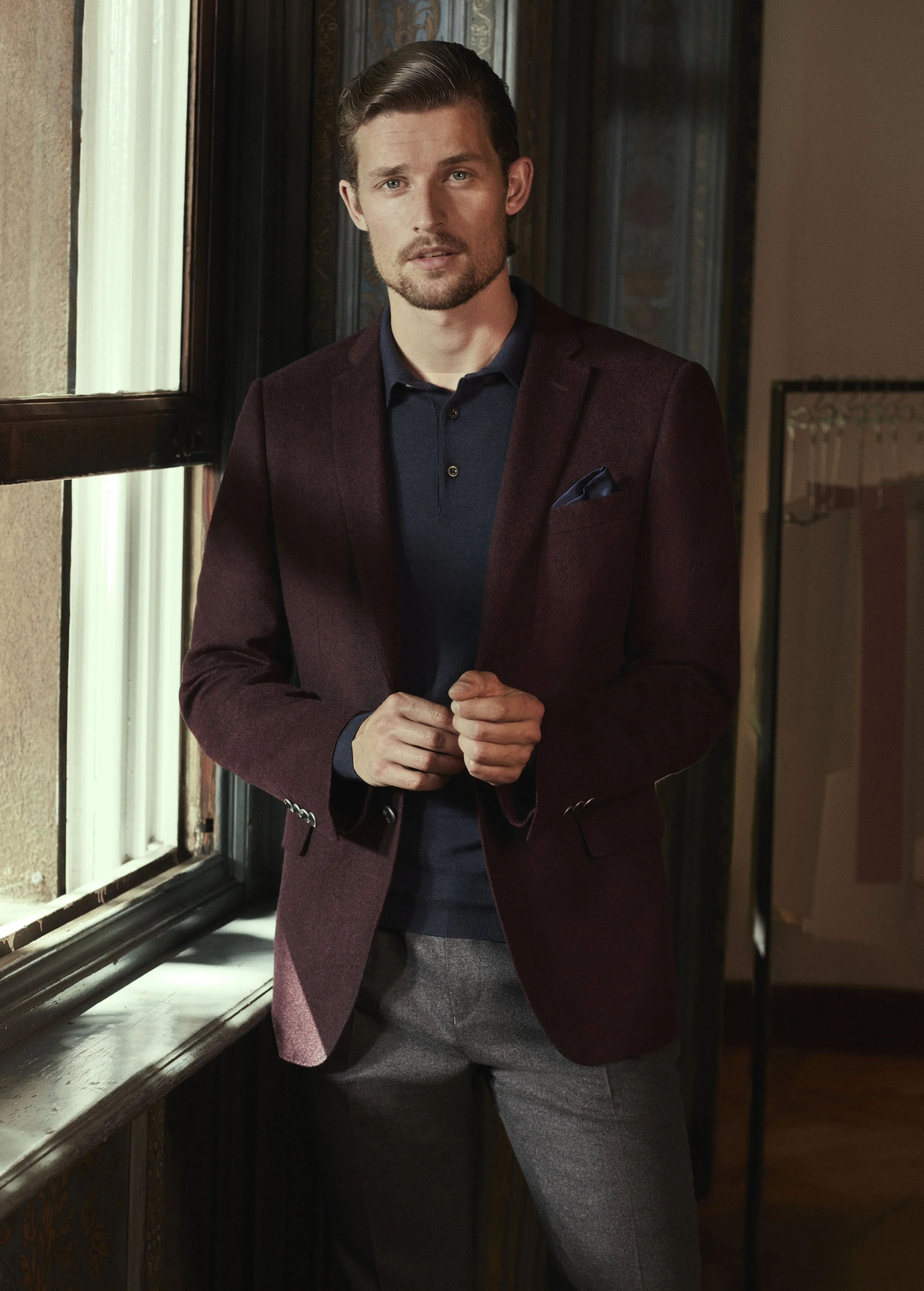 A man standing inside near a window, wearing a maroon blazer, navy shirt, and gray pants.