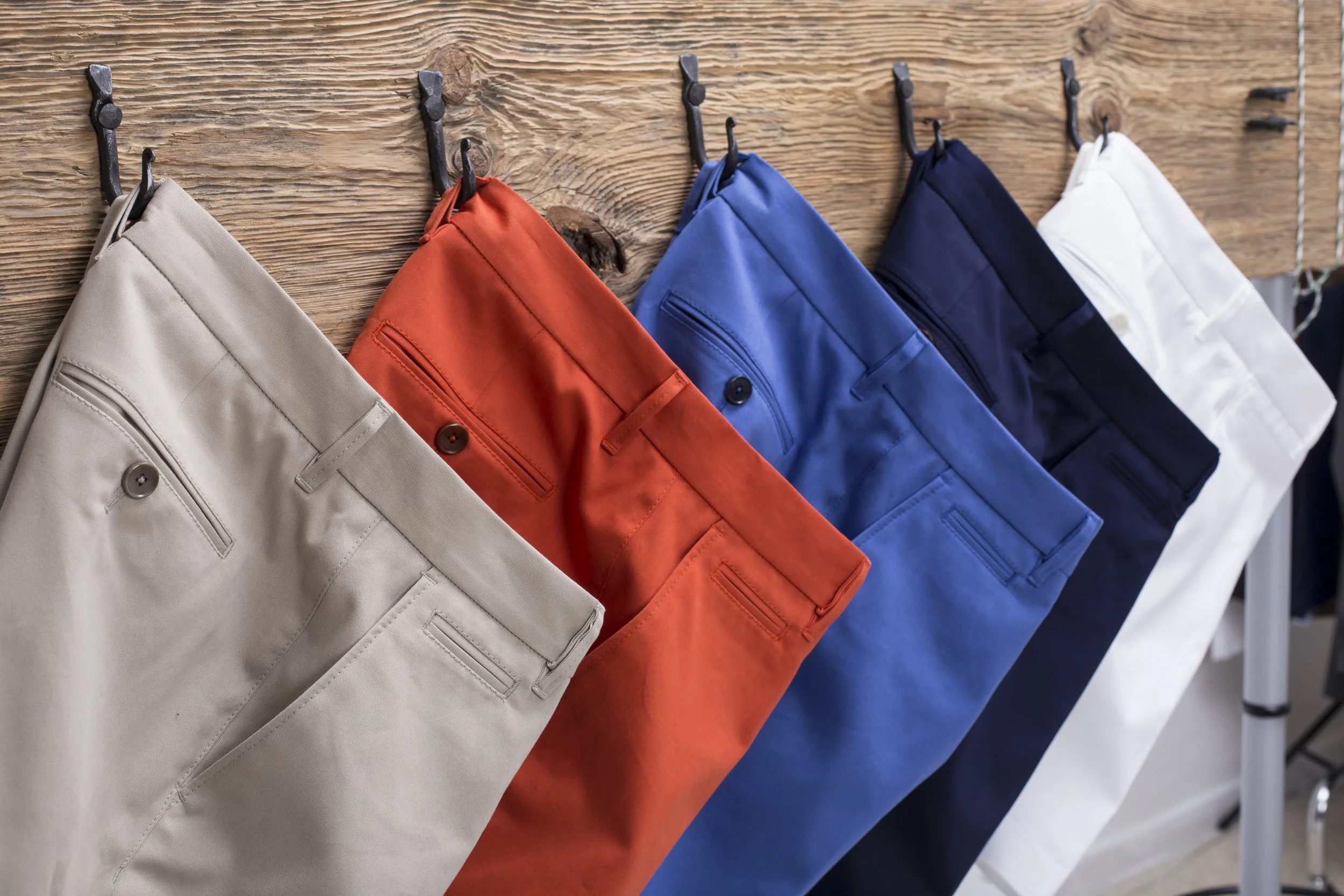 Five pairs of colorful men's pants hanging on hooks on a wooden wall, arranged in beige, orange, blue, navy, and white.