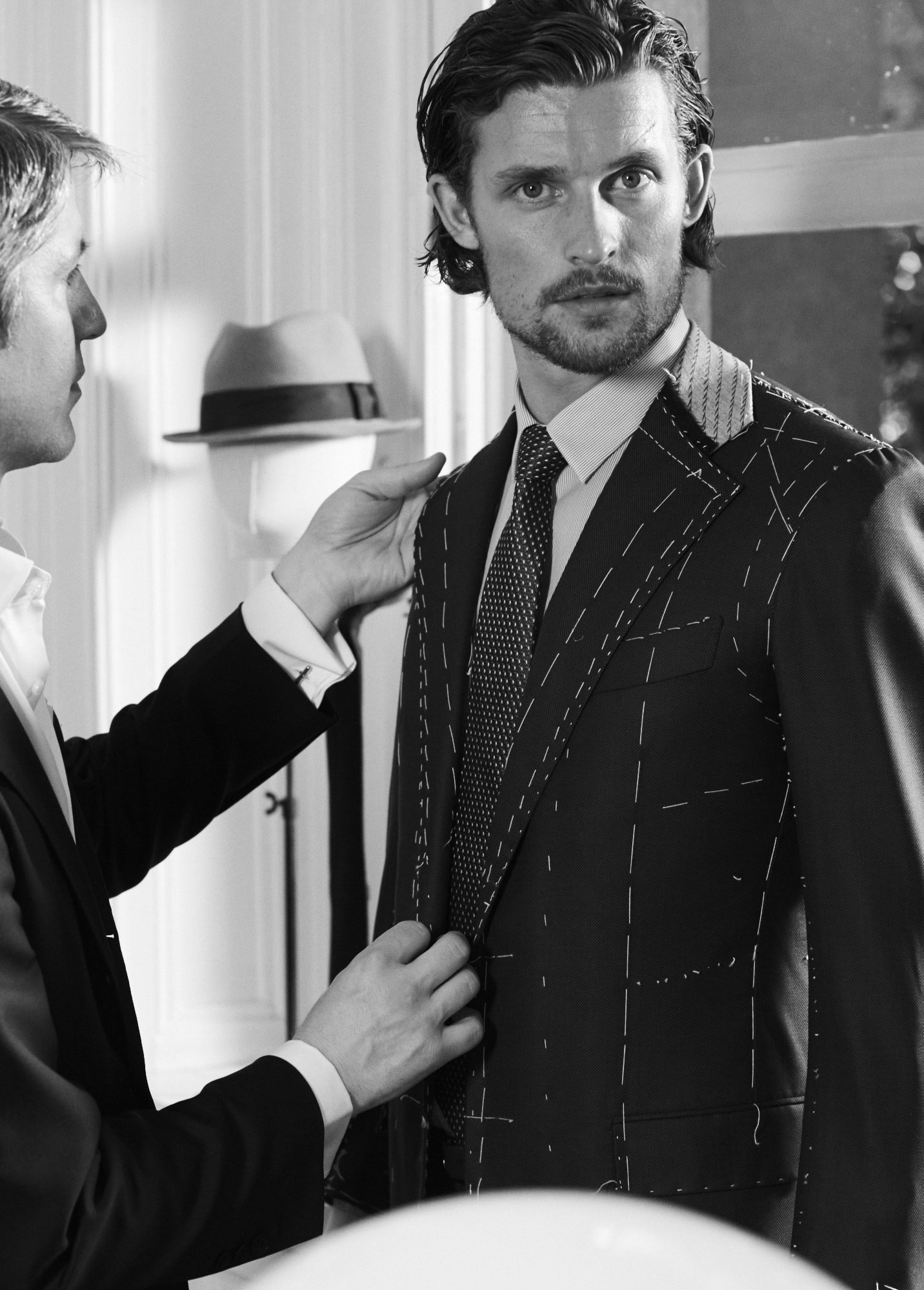 Black and white photo of a man in a basted suit being fitted for a tailored suit, with markings on the fabric indicating tailoring adjustments. Another person is pinning the suit, and a hat can be seen hanging on the wall in the background.