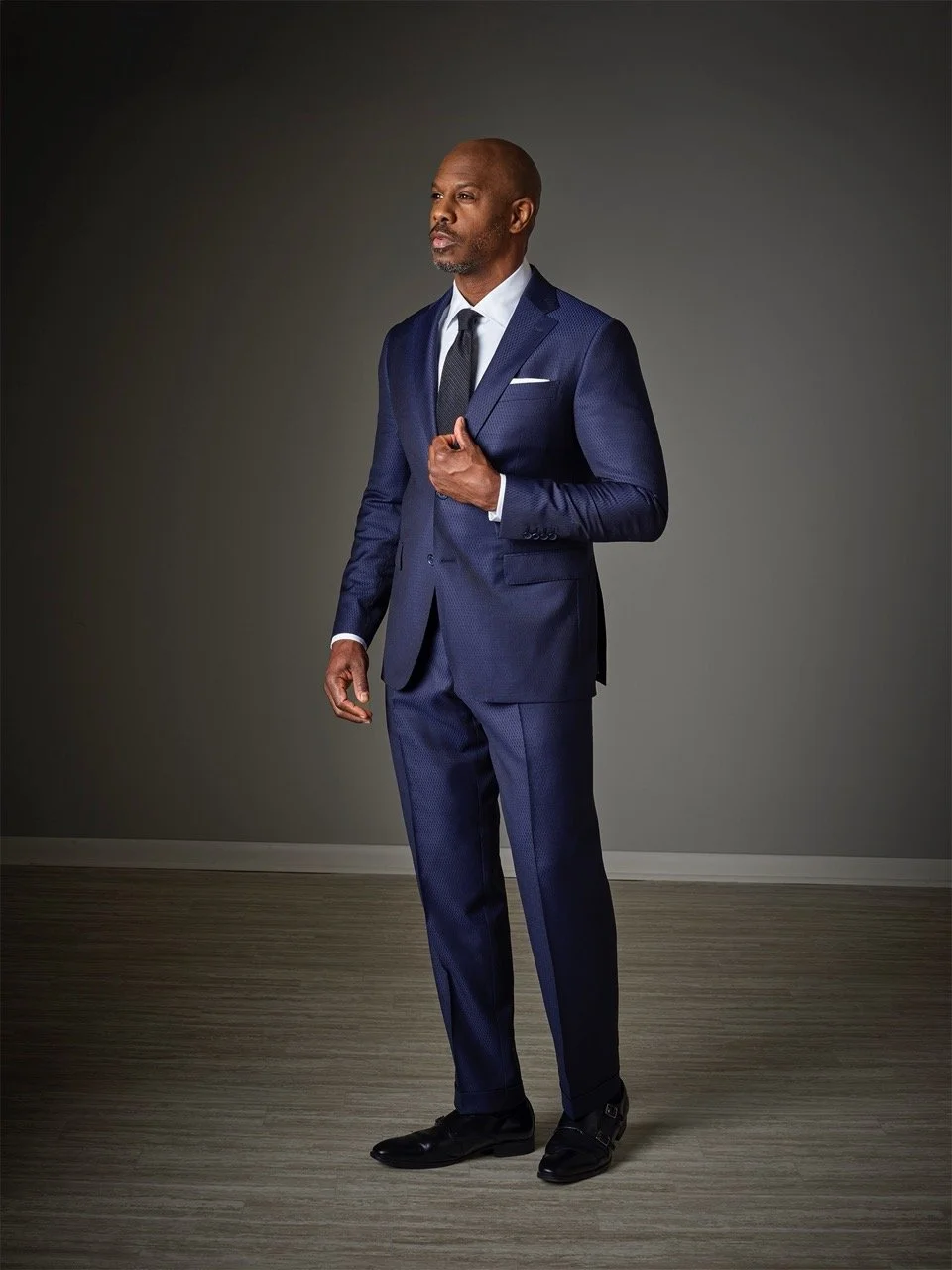 A well-dressed man in a navy blue suit standing against a plain background.