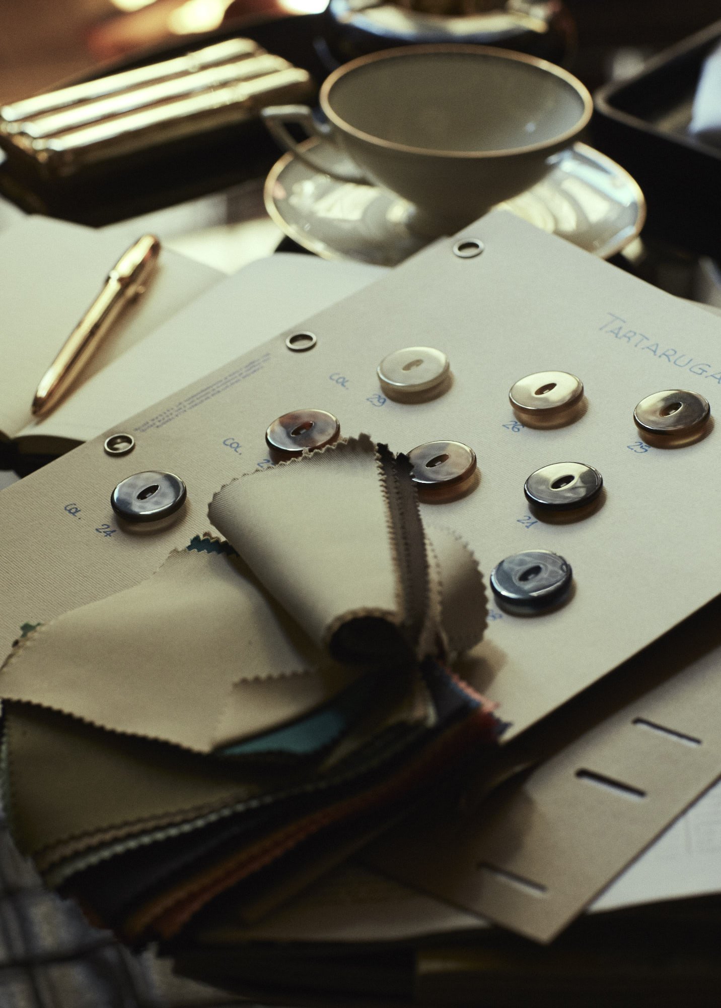 Samples of fabric swatches, buttons, and pattern sheets for color and design selection, with a tea cup and open notebook nearby on a table.
