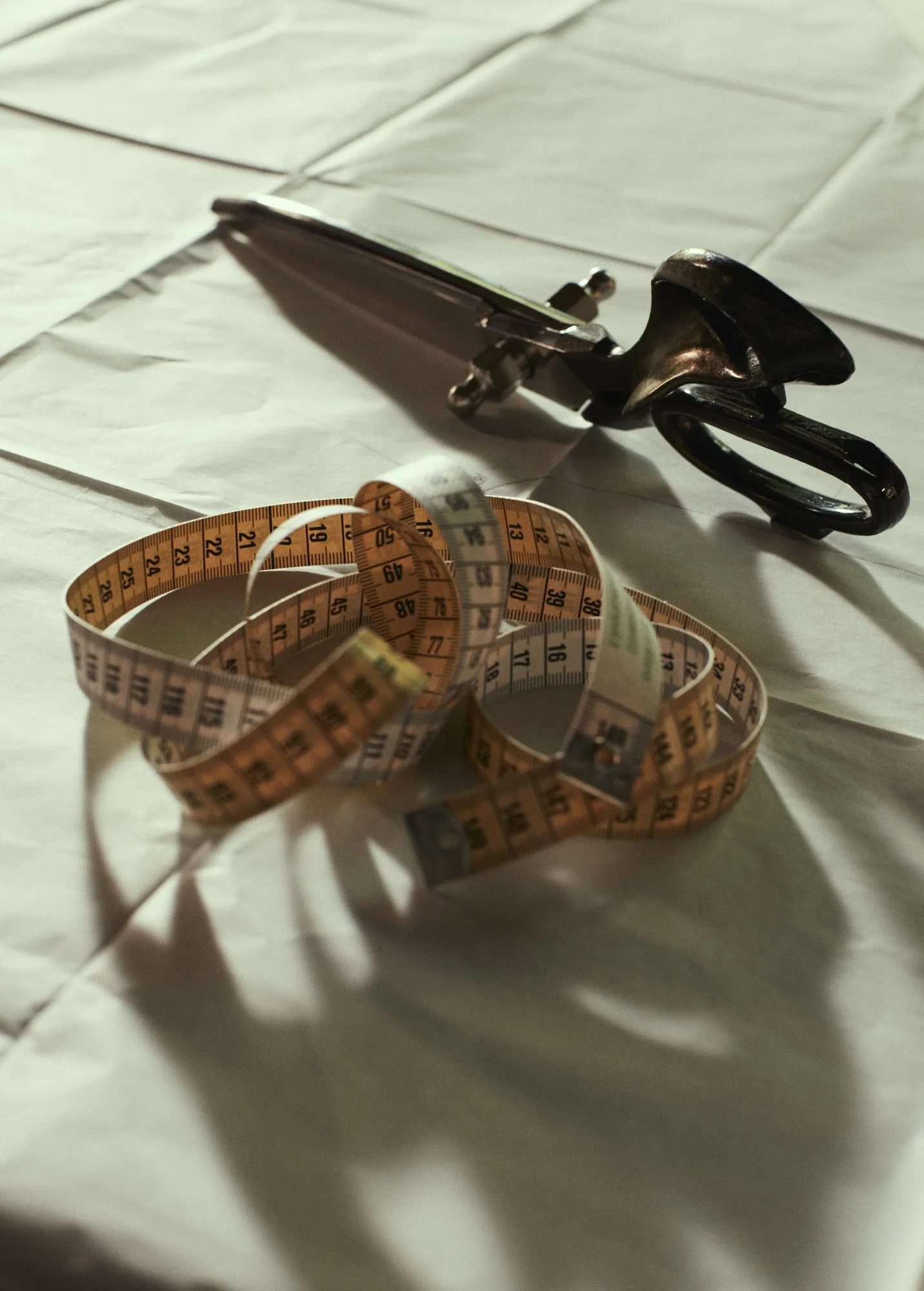 A set of shears resting on a white sheet with a yellow measuring tape loosely coiled in front of it.