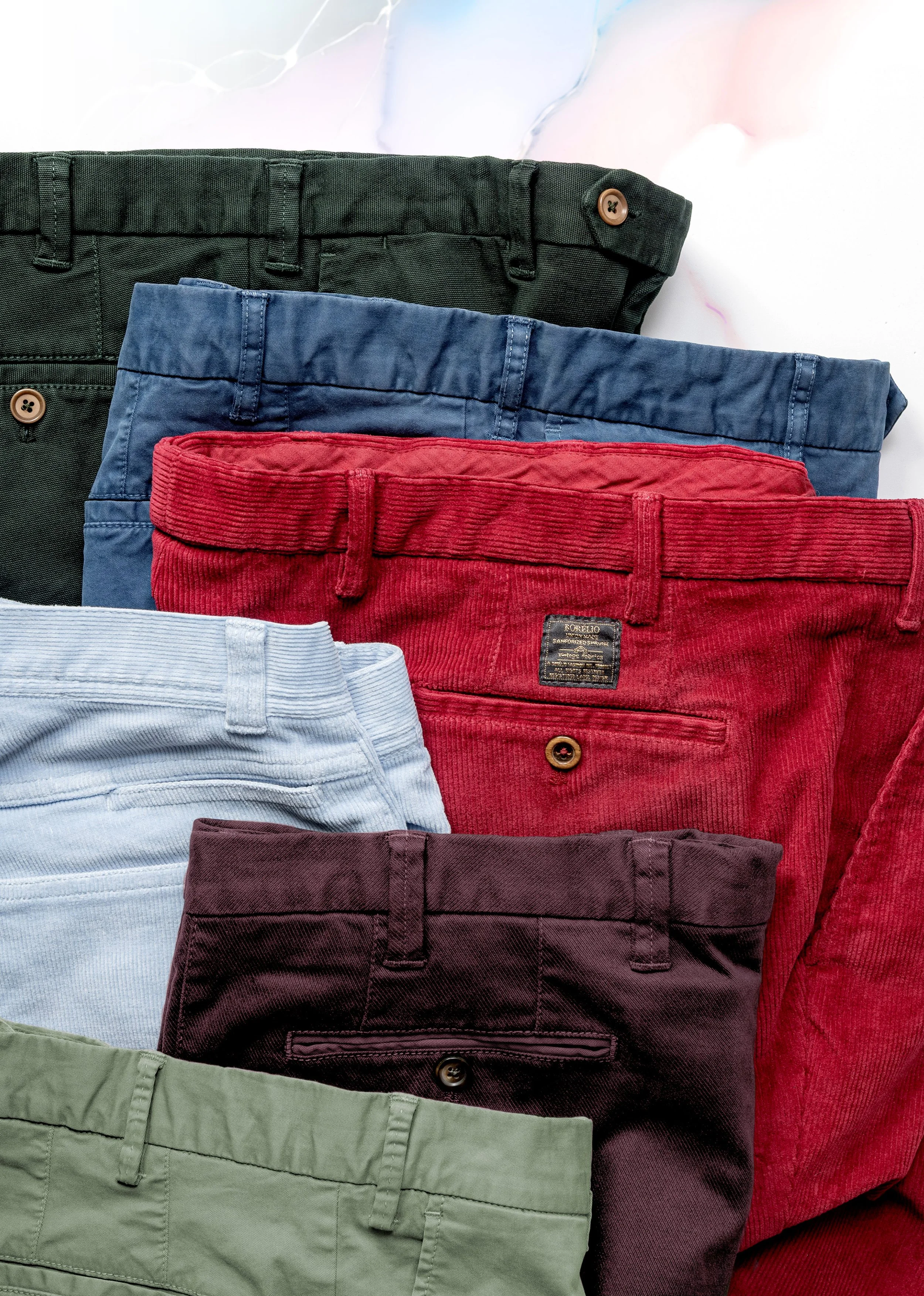 Collection of colored pants, including green, navy blue, red, light blue, burgundy, and olive green, arranged overlapping on a white background.
