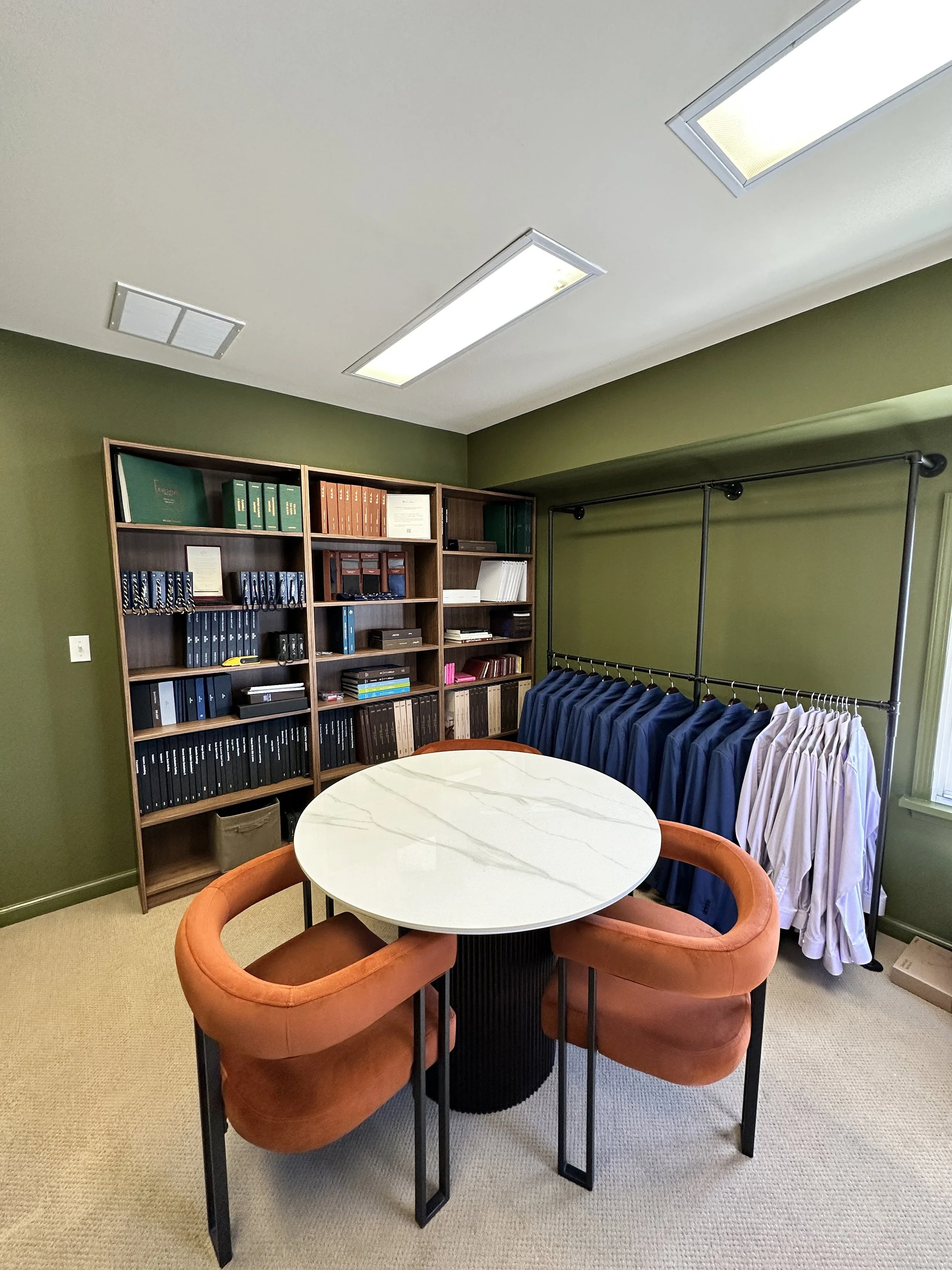 An office or conference room with green walls, a round white marble table, four orange velvet chairs, a bookshelf filled with binders and fabric swatch books, and a clothing rack with suits and dresses.