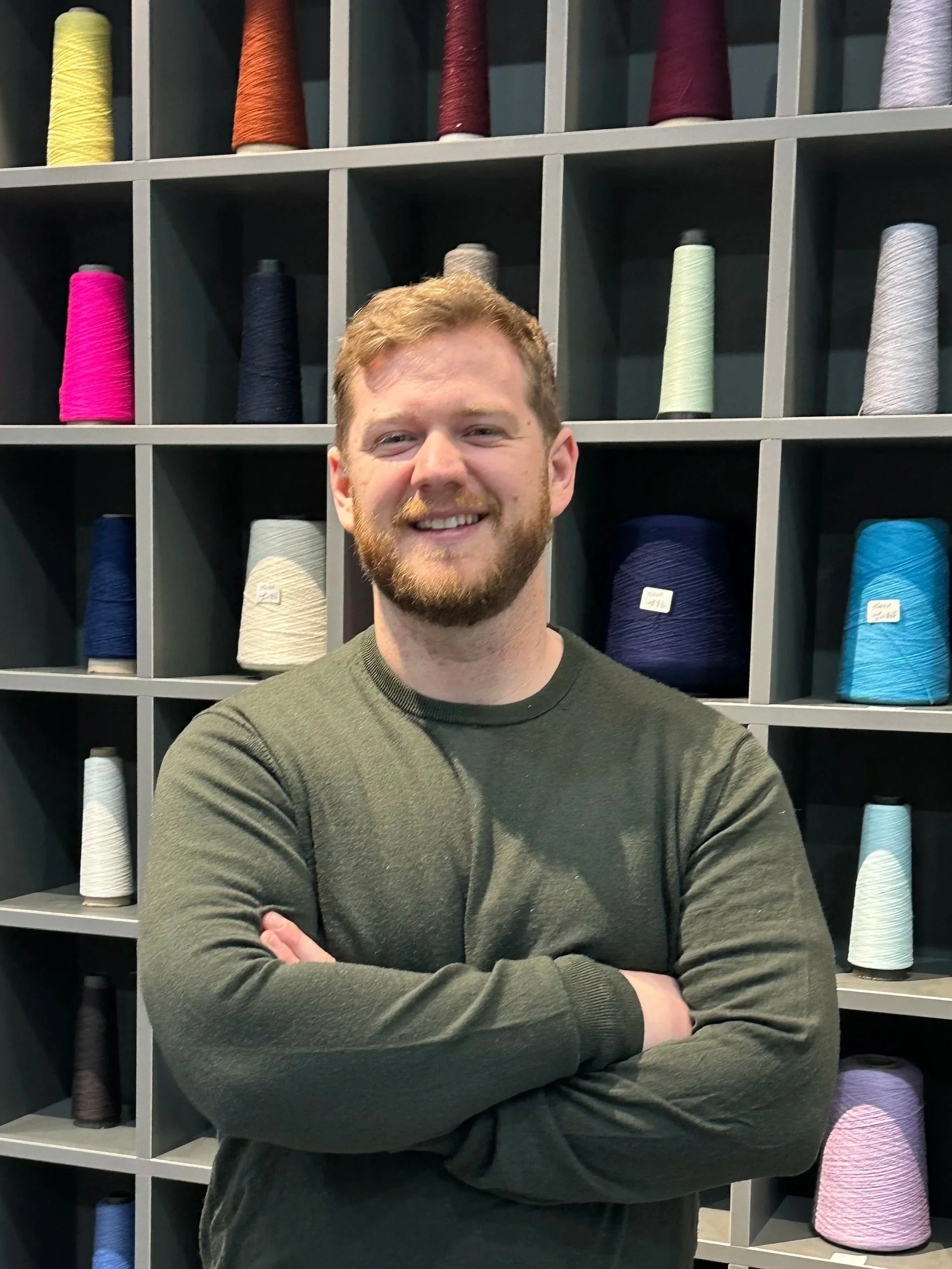 A smiling Joshua Stephen Owner and Founder with a beard and short hair standing with crossed arms in front of a  factory display wall of colorful yarn spools.
