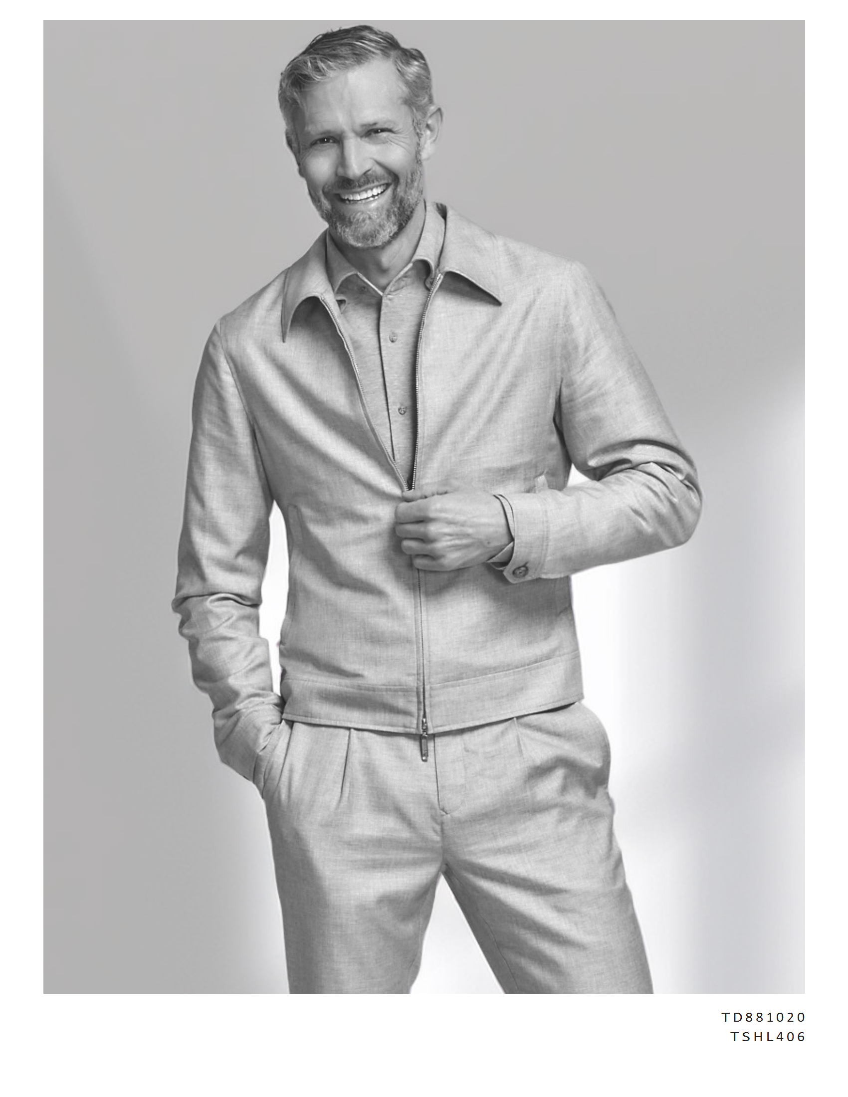 A smiling man with a beard and short hair, wearing a light-colored zip-up jacket over a button-down shirt, with one hand in his pocket and the other adjusting his jacket, against a plain background.